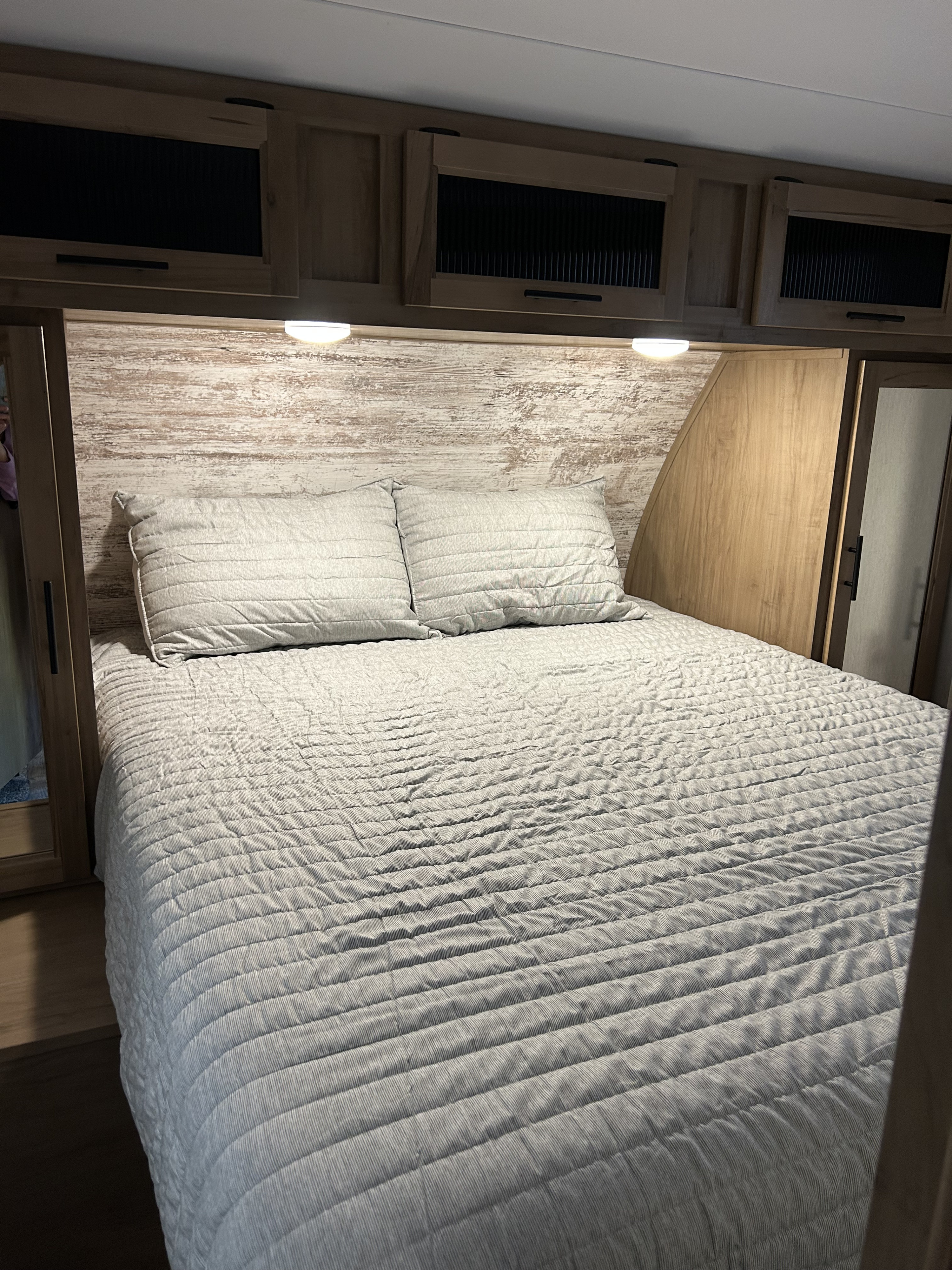 Cozy RV rental bedroom featuring a bed with gray bedding, a distressed wood headboard, and overhead storage.. Forest River Cherokee Arctic Wolf 2023