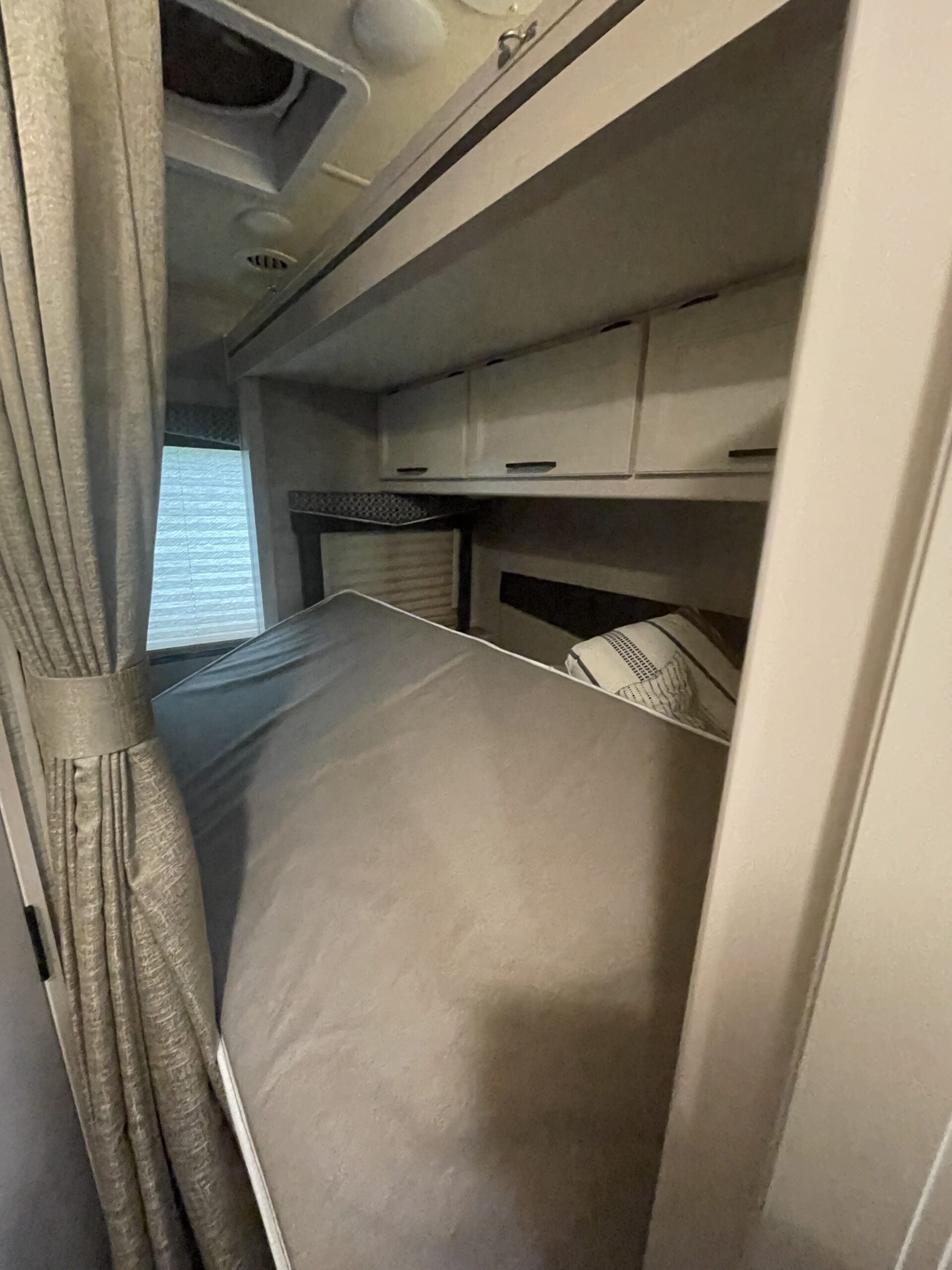 An RV rental bedroom with a large mattress, patterned curtains, window blinds, and practical overhead storage cabinets.. Thor Chateau 2021