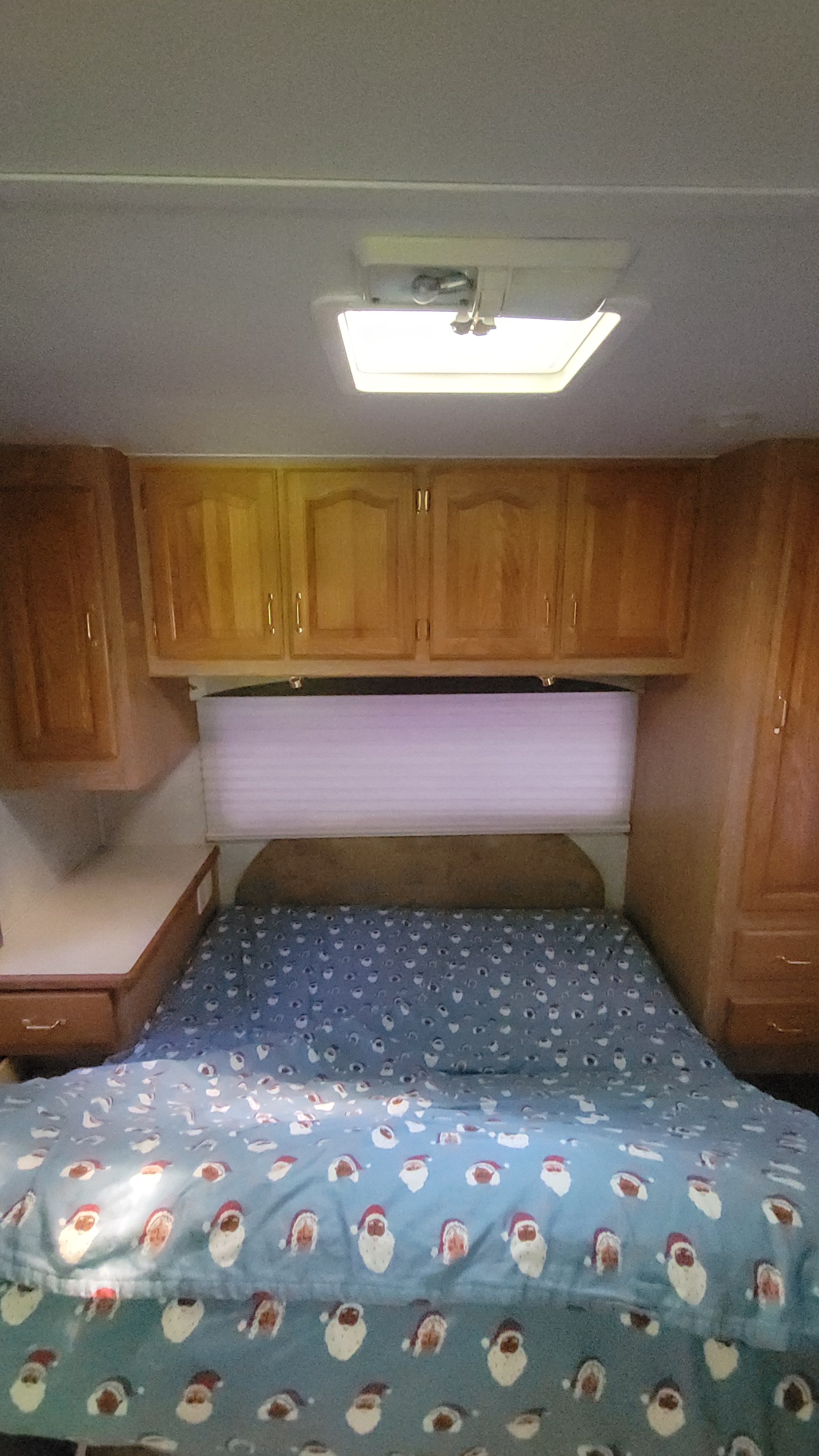 Inside an RV rental: a compact bedroom with a fun Santa blanket, wood cabinets, and bright overhead lighting.. Forest River Sunseeker 2003