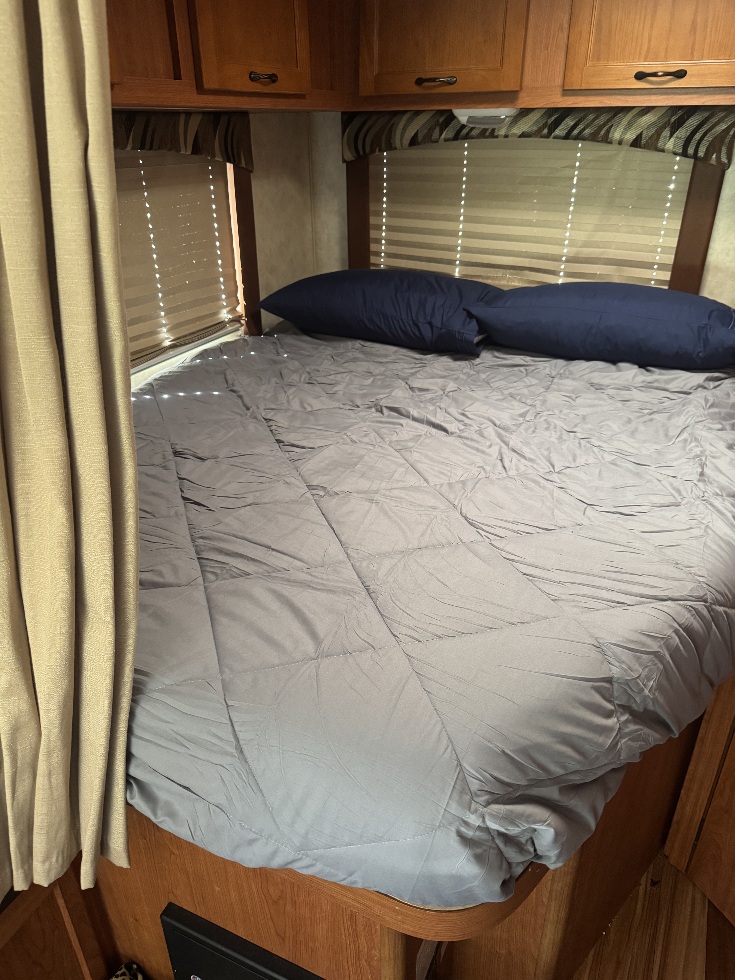 Cozy rv rental bedroom: grey comforter, navy pillows, wooden cabinets, sunlit windows, and beige curtain.. Coachman Freelander 21QB Chevy 4500 2013