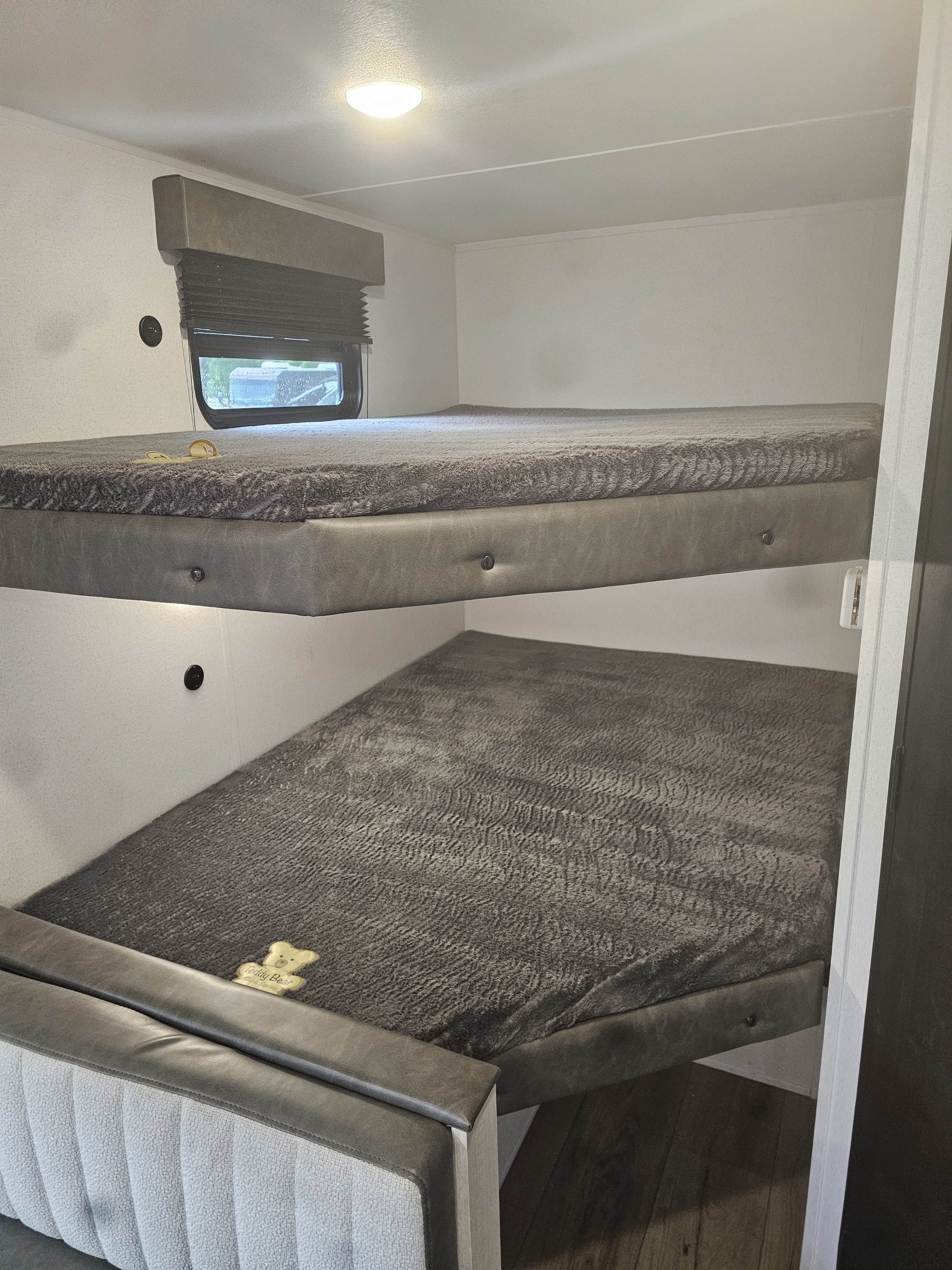 RV rental bunk beds feature soft grey bedding, a window with a blind, and overhead lighting. A cozy sleeping area for family travel.. Forest River Palomino Puma Camping Edition 2026