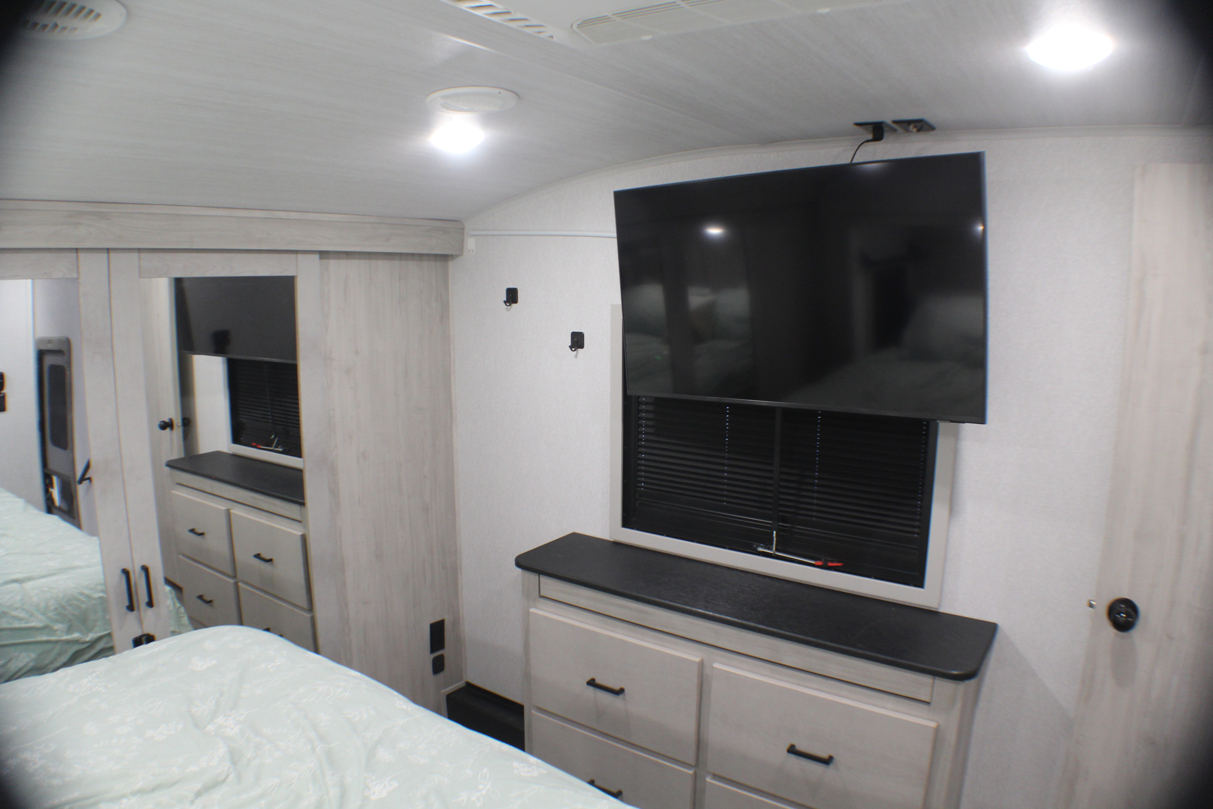 Comfortable RV rental bedroom featuring a bed, mirrored wardrobe, large TV mounted above window blinds, and storage dresser.. East to West Blackthorn 2025