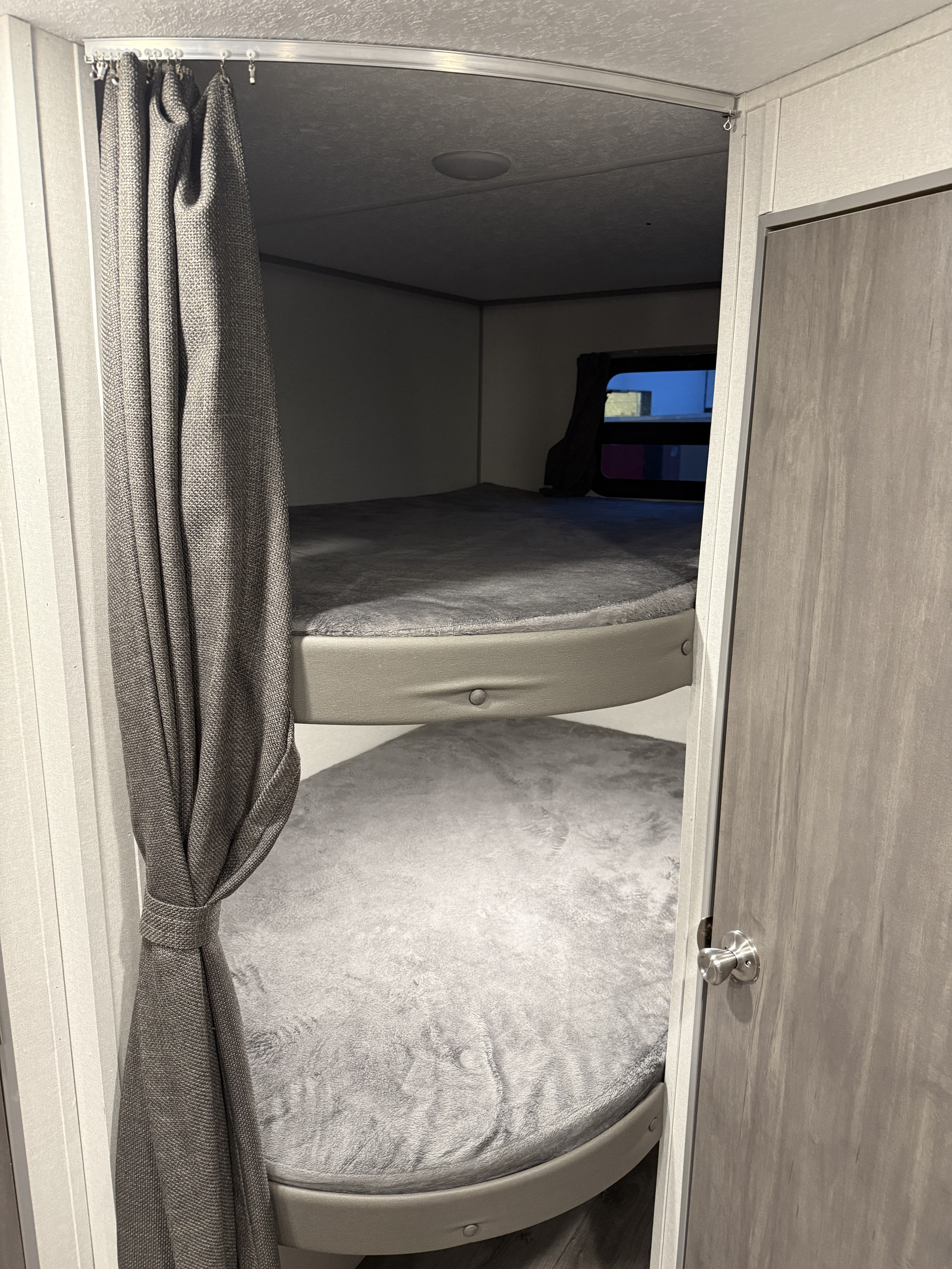 Cozy RV rental bunk beds with soft gray bedding and a privacy curtain, offering a compact sleeping area for family travel.. Forest River Coachmen Catalina Summit 2025