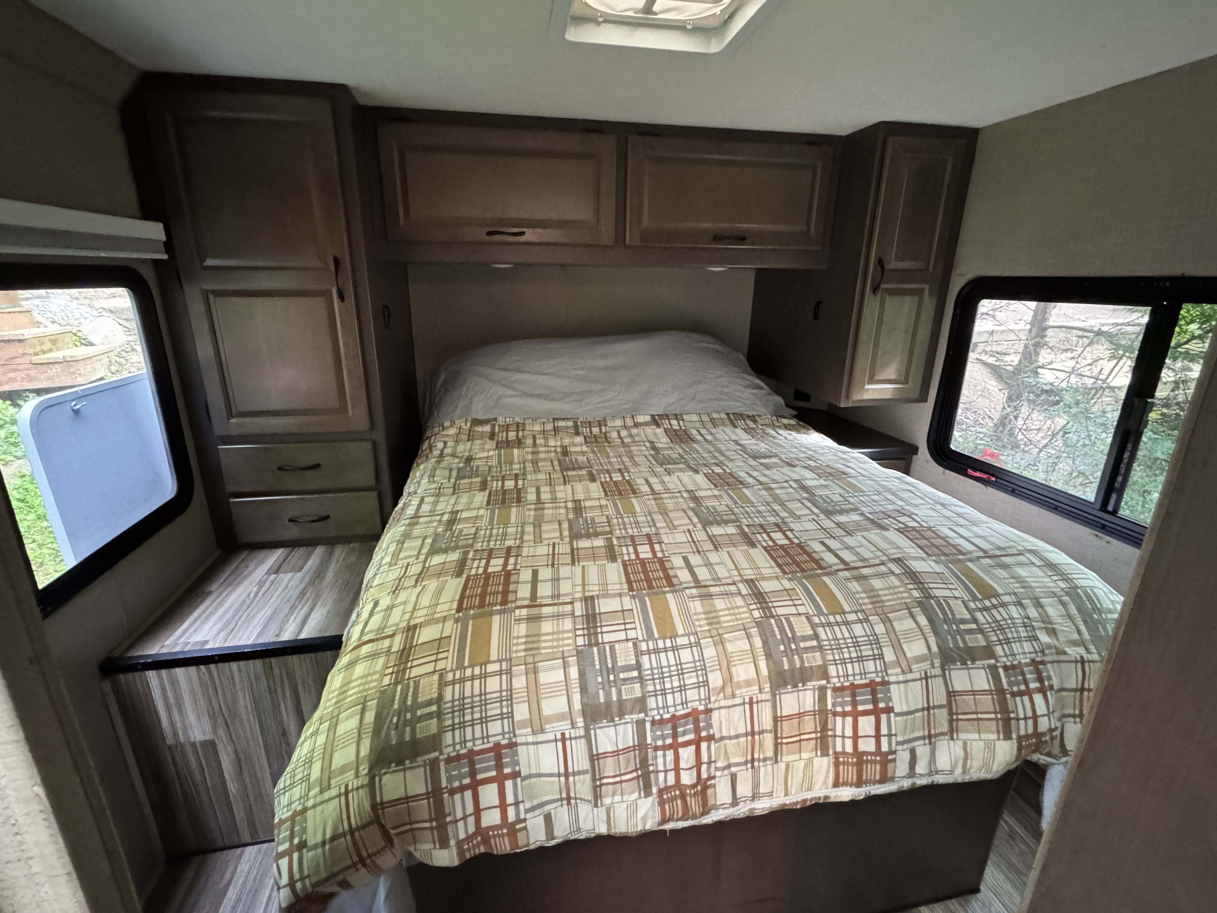 RV rental bedroom: a bed with plaid comforter, wood cabinets, and windows showing outdoor trees and stairs. Compact and cozy.. Thor Four Winds 2018