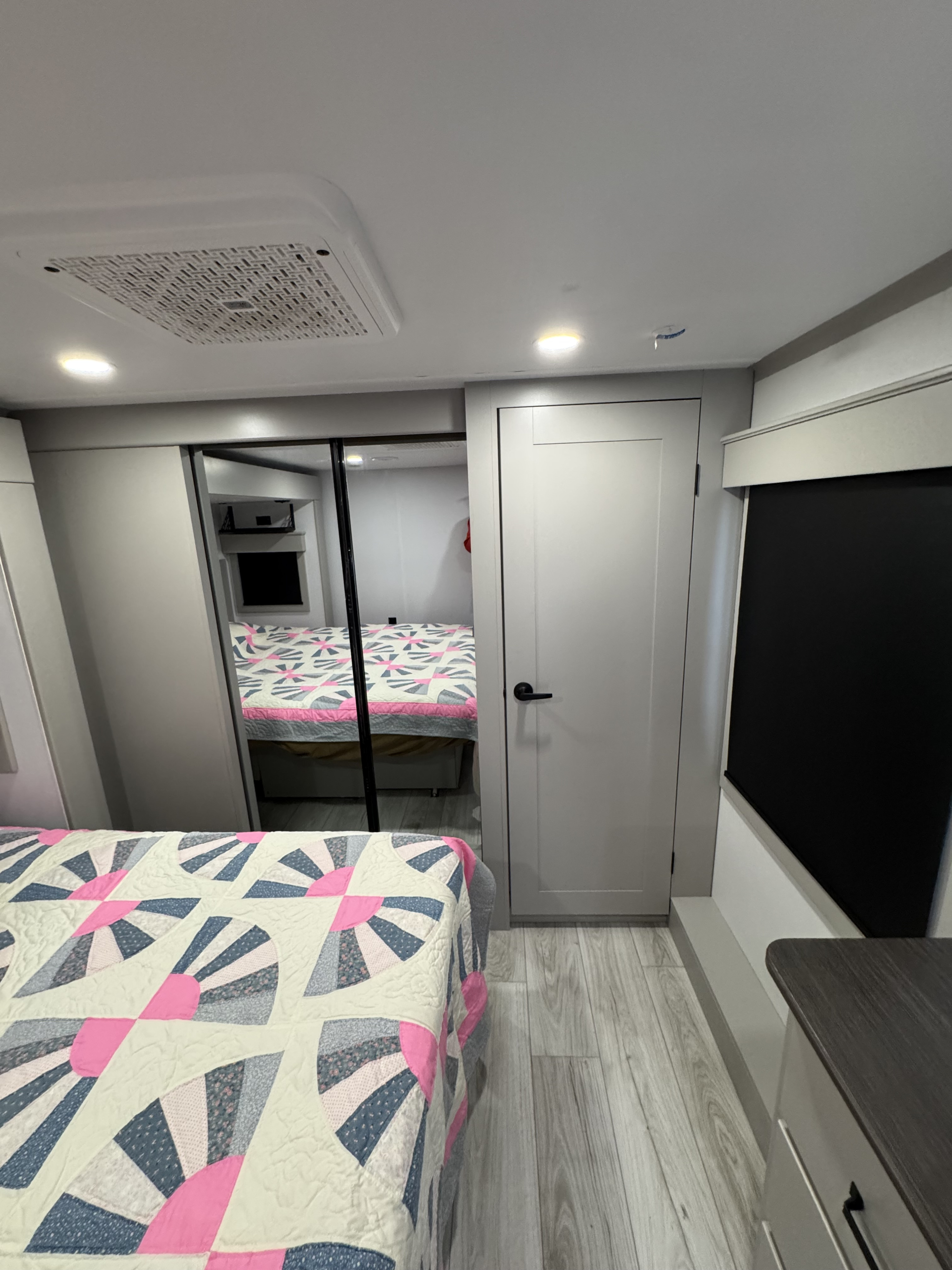 Compact RV rental bedroom featuring a colorful quilt, mirrored closet reflecting the bed, recessed lights, and gray wood floor.. Keystone RV Montana 2025