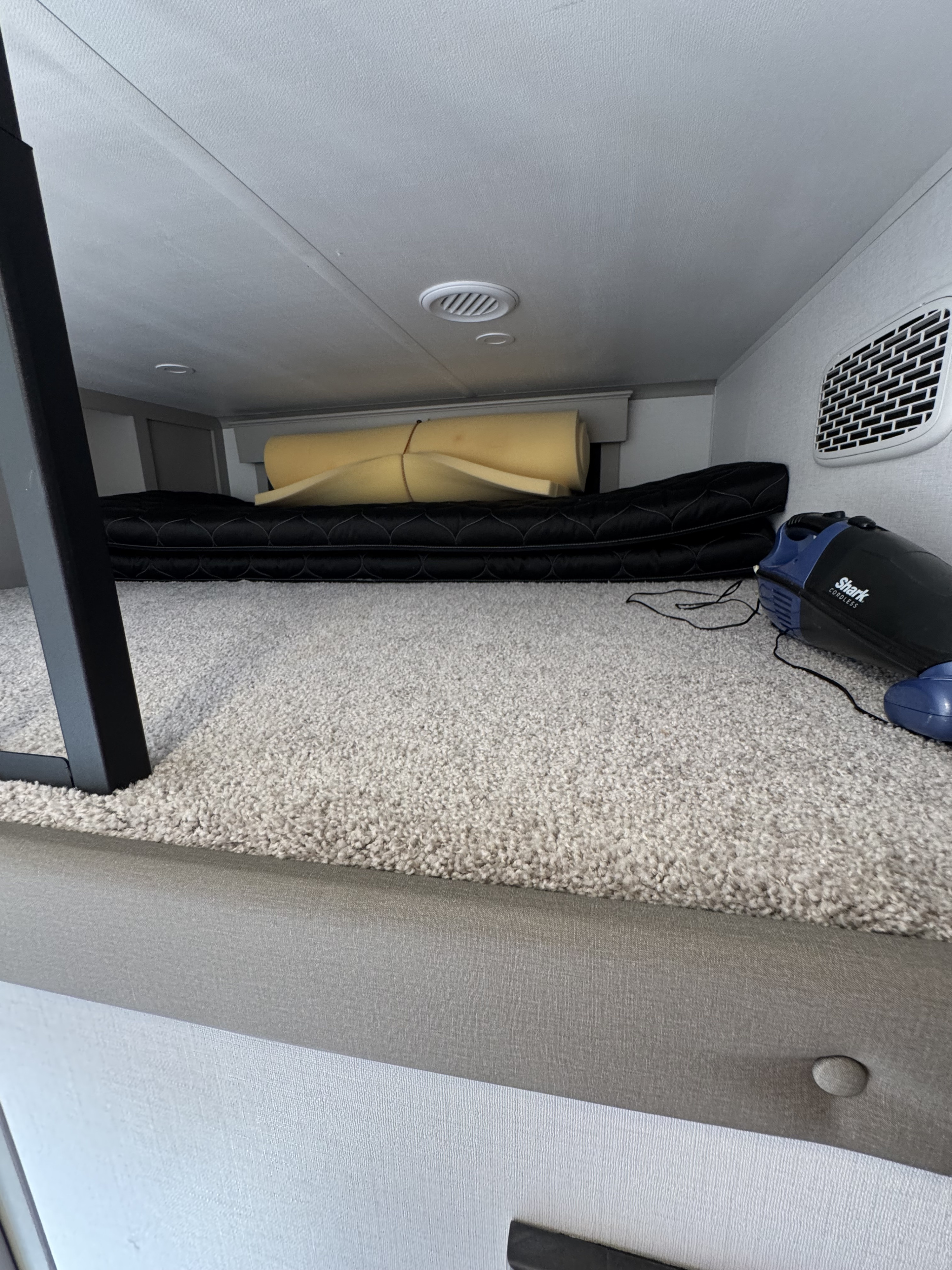 RV rental bunk with a black quilted mattress, stored yellow foam pad, and a Shark handheld vacuum on light beige carpet.. Keystone RV Montana 2025