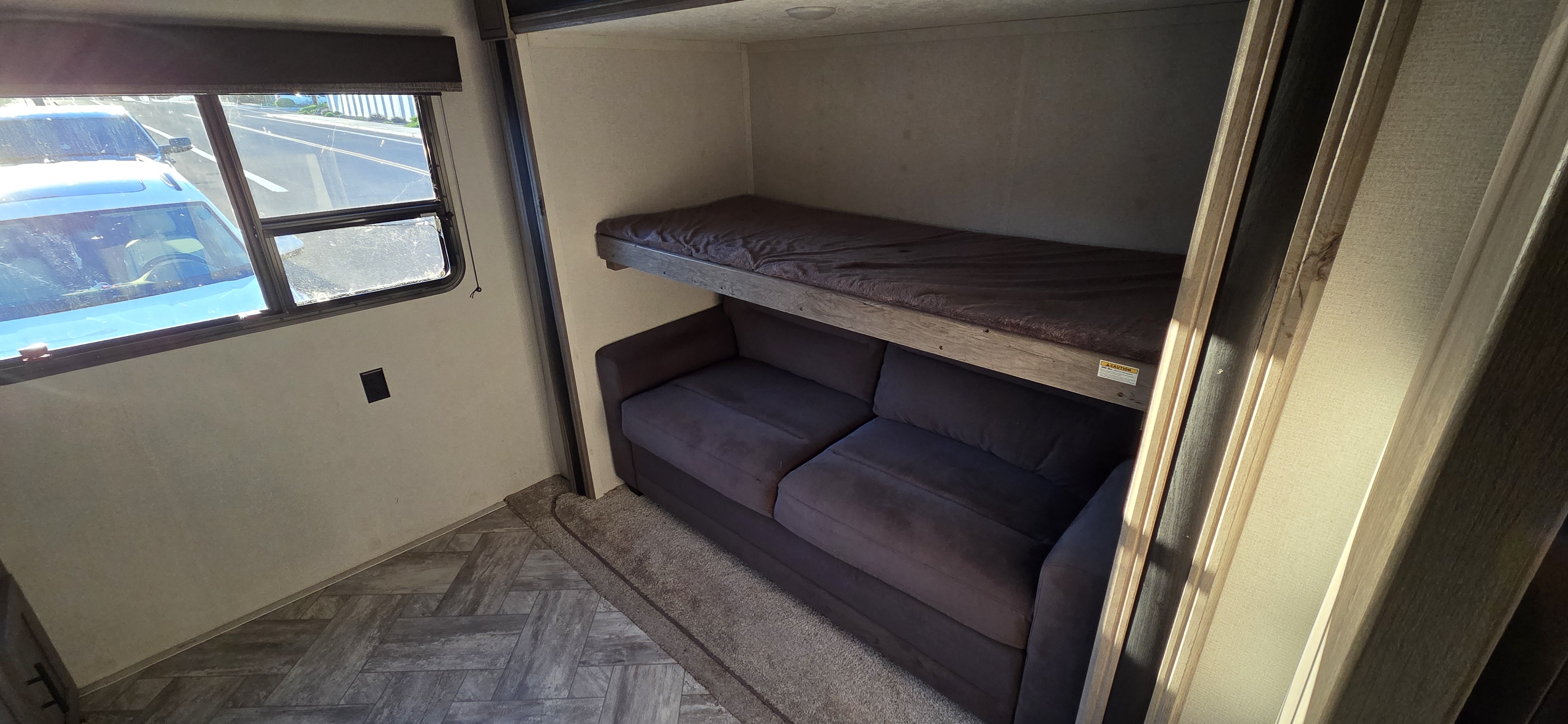 This RV rental interior features a cozy bunk bed above a dark sofa, beside a window with sunlight, and a patterned floor.. Keystone RV Springdale 2021