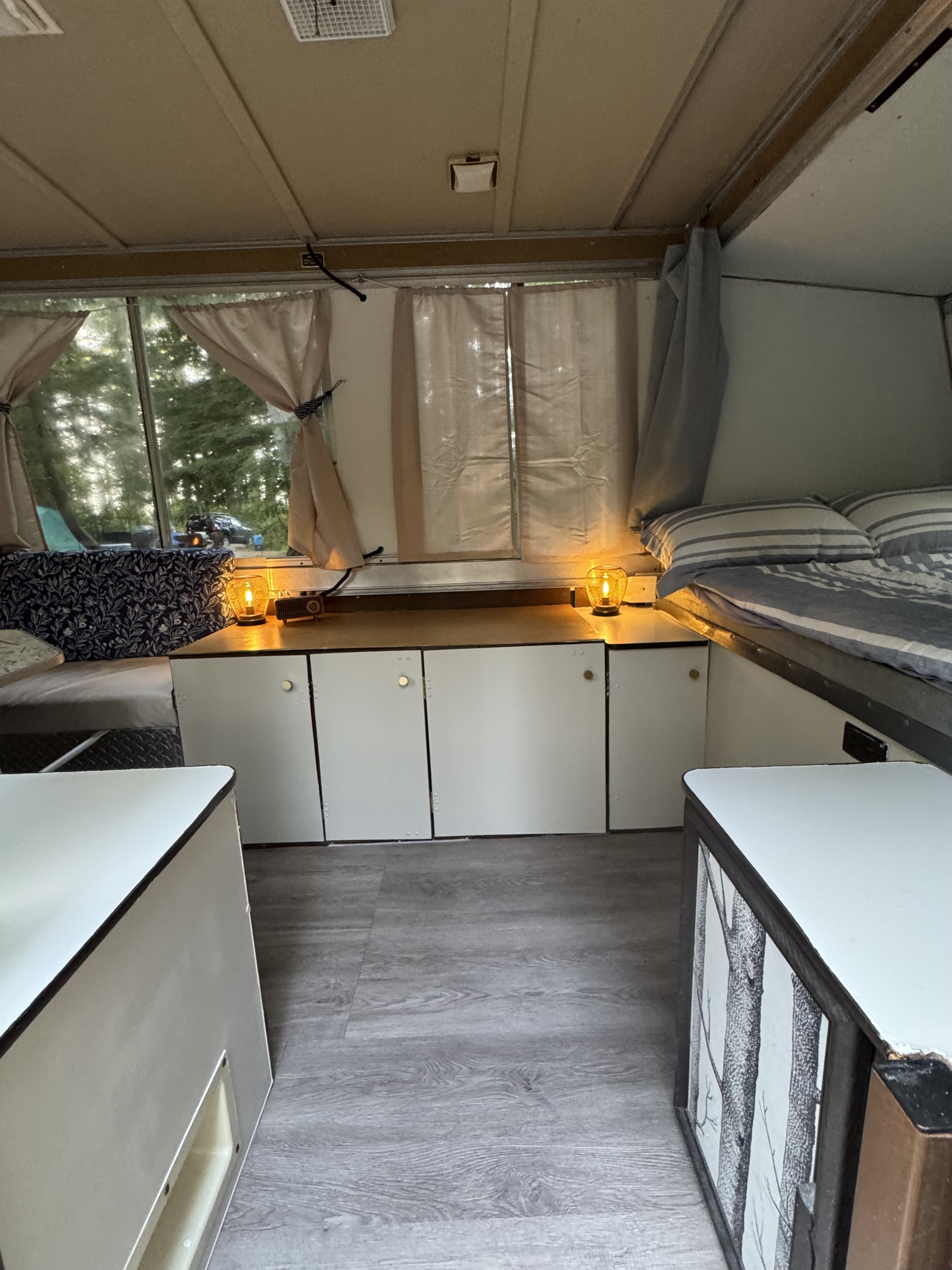 Pop-up RV rental interior with cozy beds, storage cabinets, and warm lamps. View of trees outside through the windows.. Apache Mesa 1977