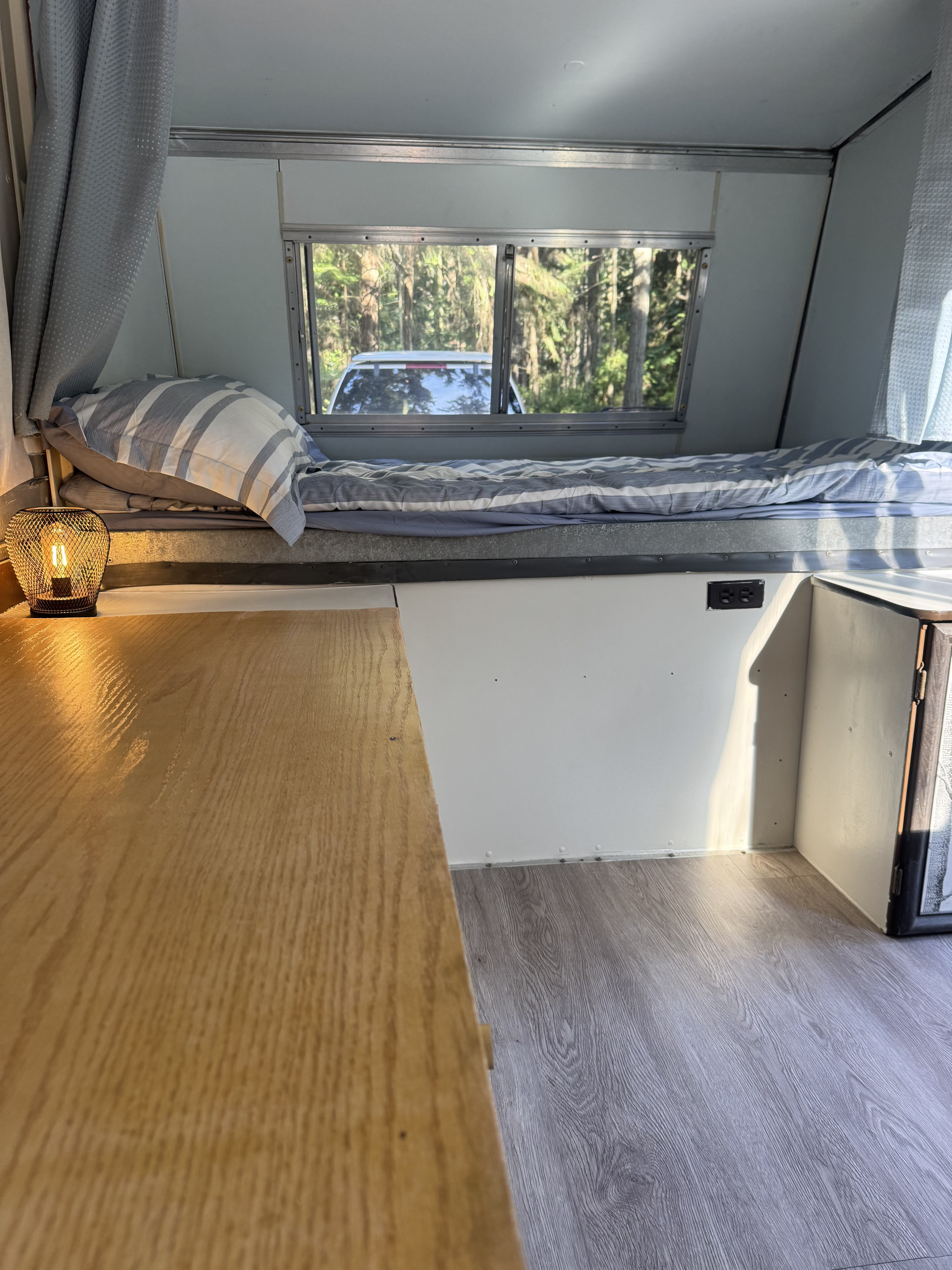 Inside a cozy RV rental: bed with striped bedding, wooden table, modern lamp, window view of trees, and gray flooring.. Apache Mesa 1977