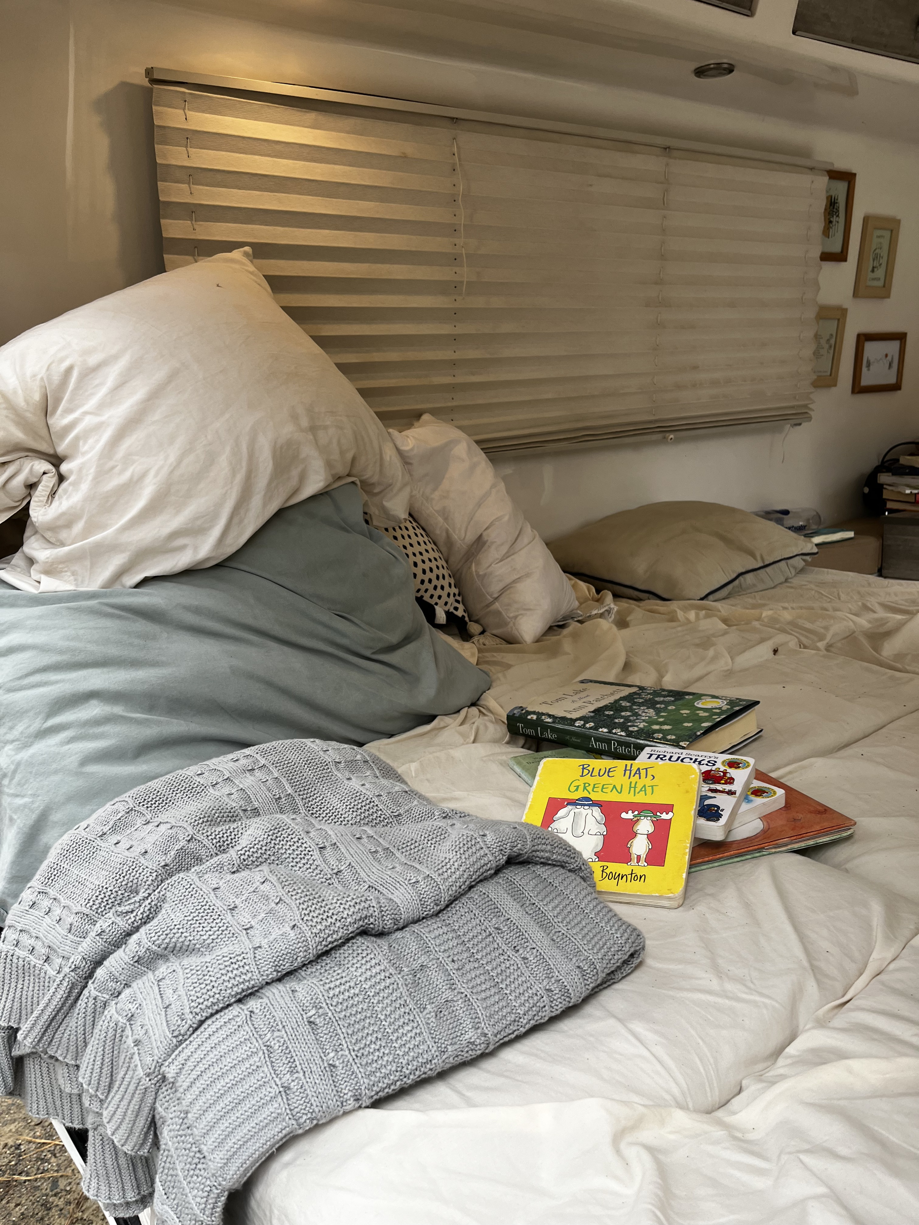 Cozy rv rental bed with white and light blue pillows, grey knitted blanket, a stack of books, and horizontal window blinds.. Happier Camper HC1 2022