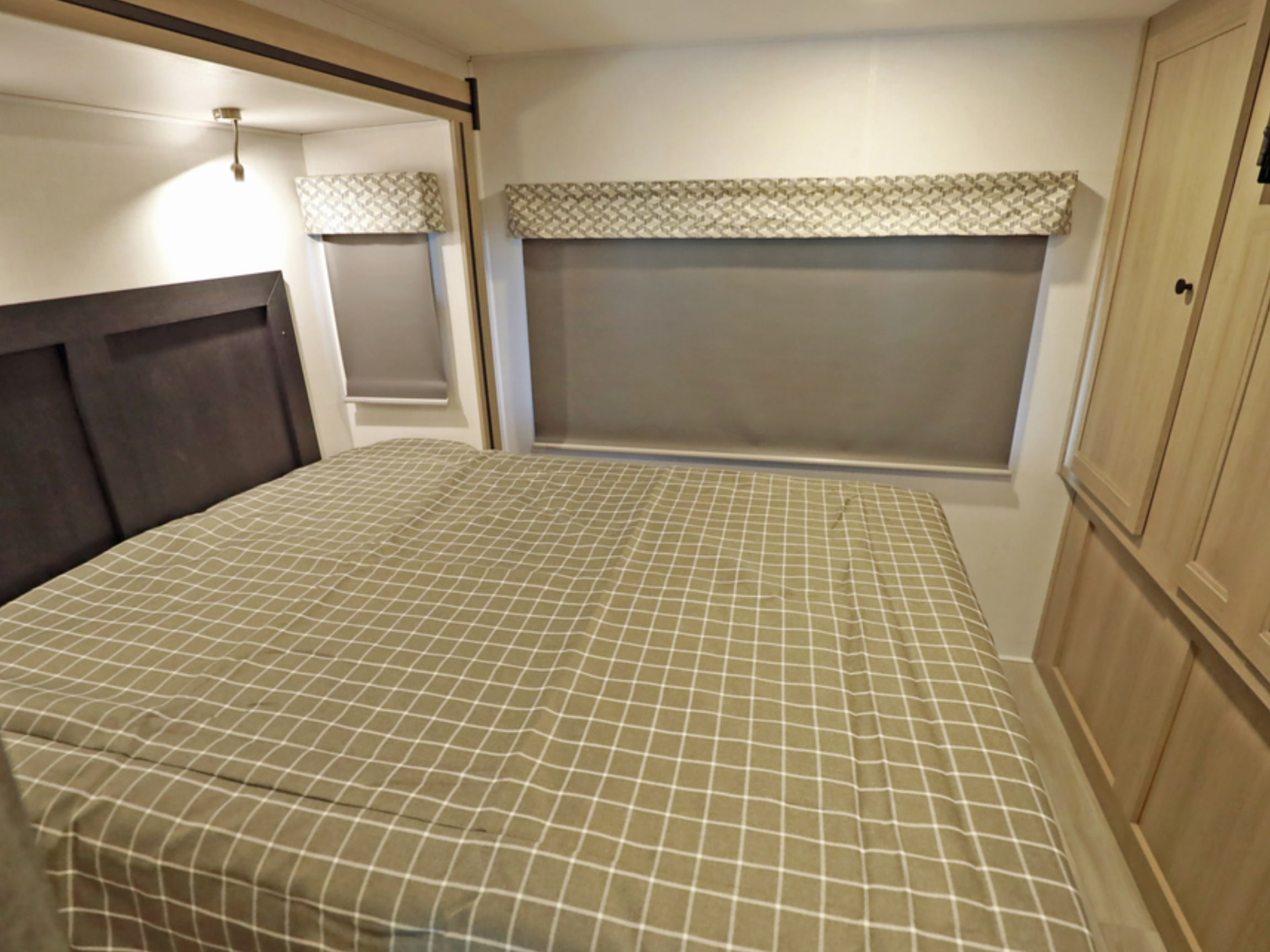 RV rental bedroom with a checkered bed, dark headboard, windows with blinds, light fixture, and wooden cabinets.. Classe C C31 pieds avec extension(s) 2023