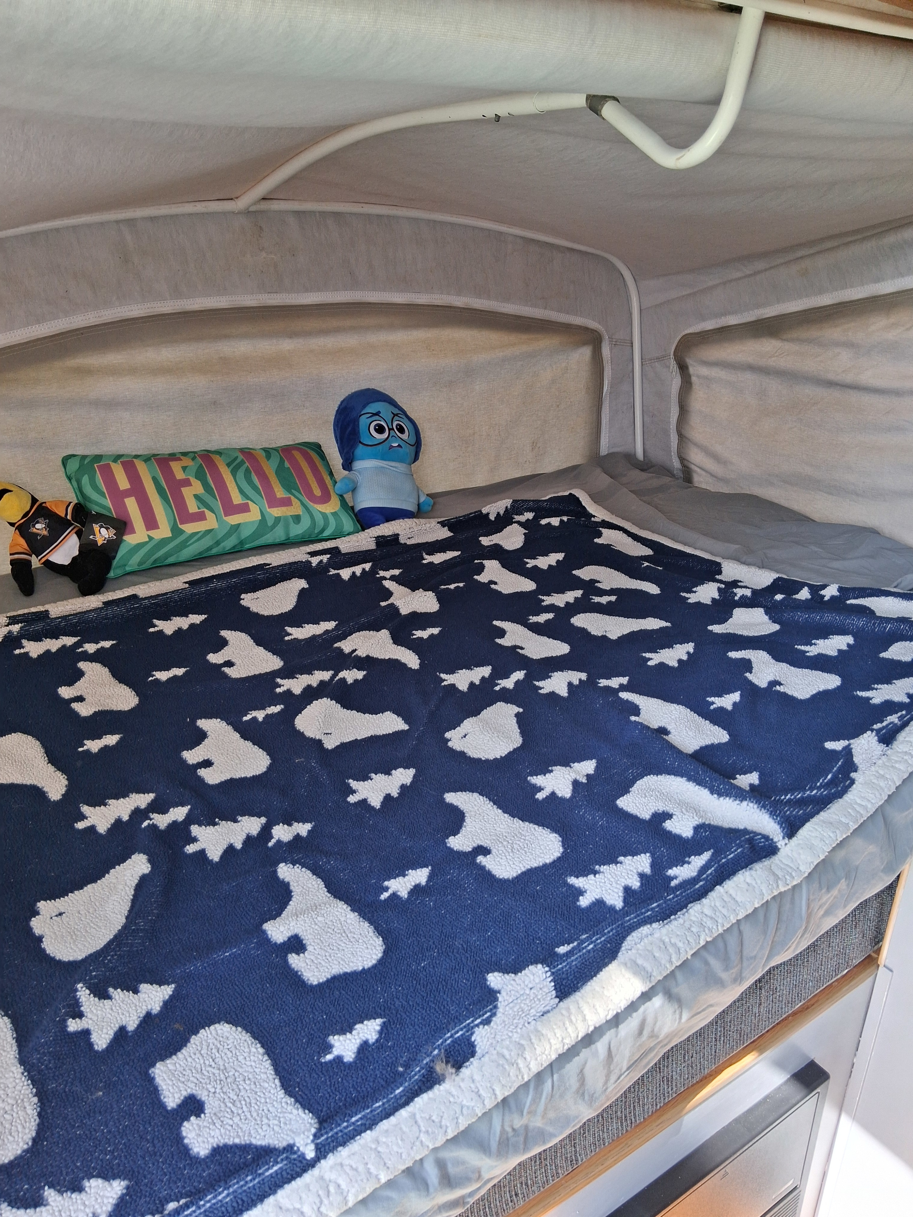 Cozy RV rental bed with a blue blanket, "Hello" pillow, and plush toys in a pop-up camper interior.. Dutchmen Kodiak Cub 2000