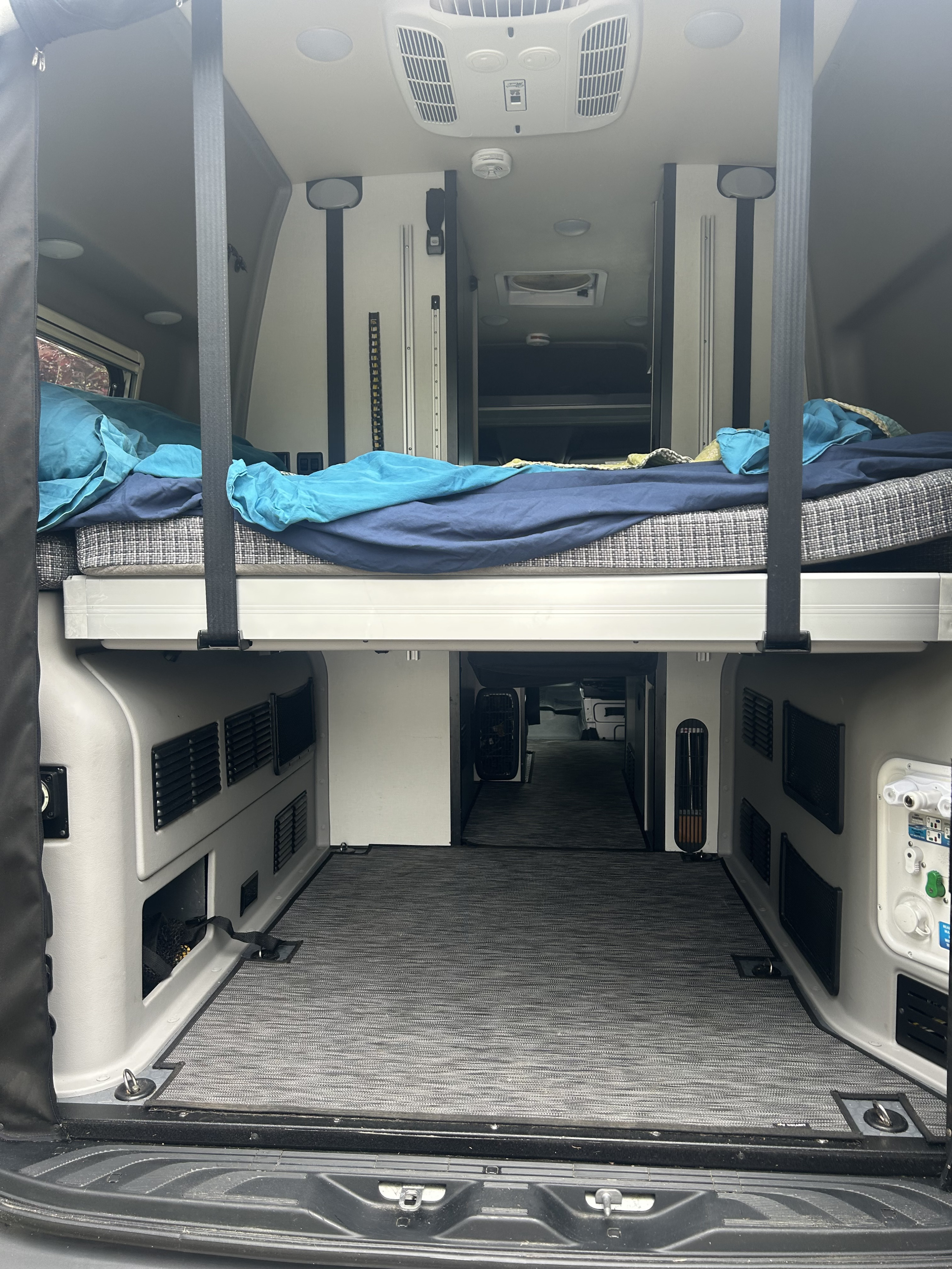 Rear interior of an rv rental camper van showcasing a raised bed with bedding, ample under-bed storage, and a well-equipped ceiling.. Mercedes-Benz Revel 2022