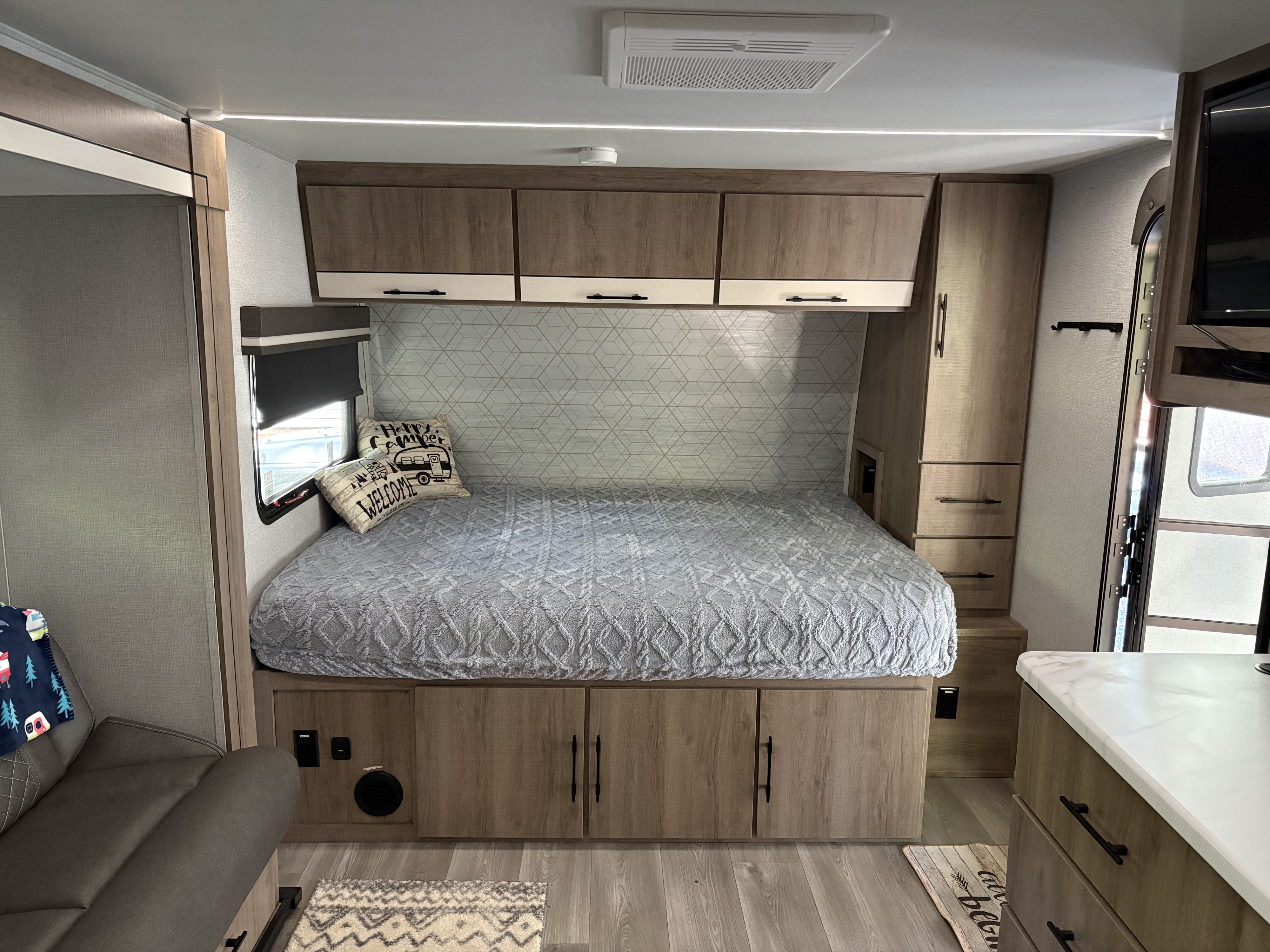 Cozy and modern RV rental bedroom featuring a comfy bed, overhead storage, patterned wall, and natural light from the window.. Grand Design Imagine AIM 2024