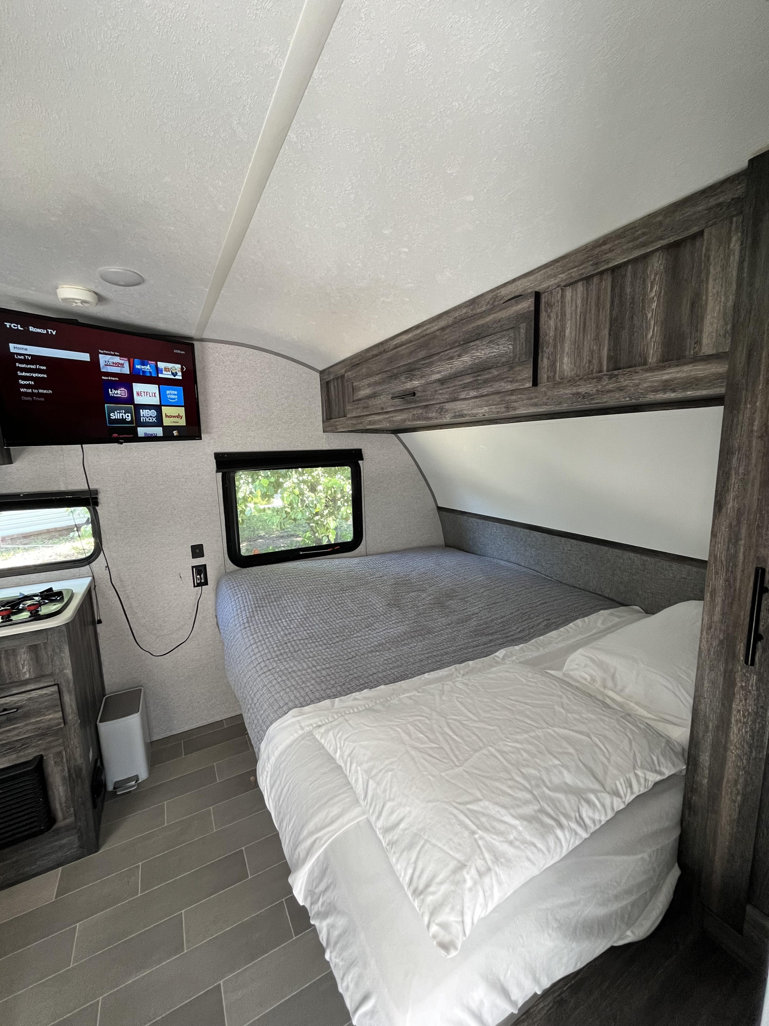 Cozy rv rental bedroom featuring a large bed, wall-mounted TV, wood cabinets, and a compact kitchen area with a stove.. Forest River Salem FSX 2021