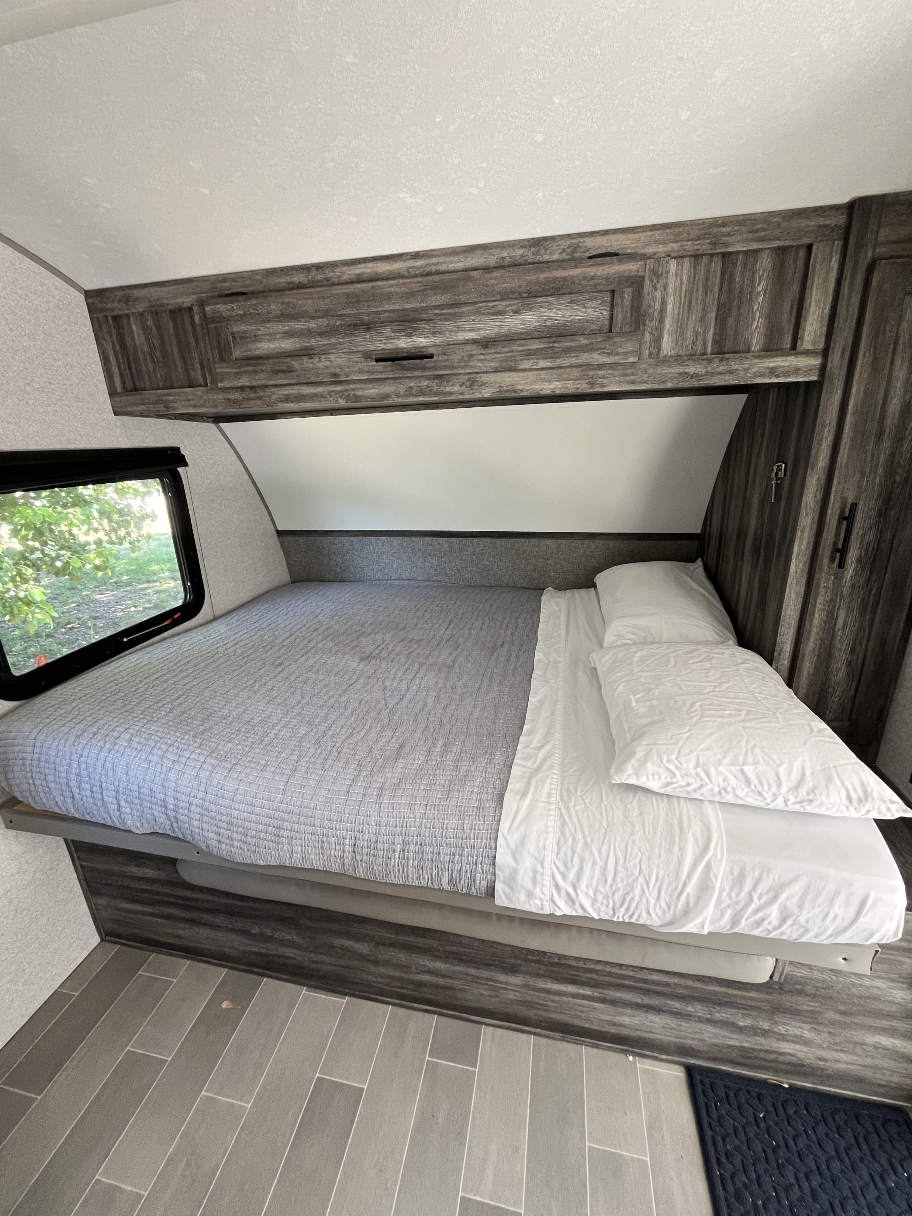 Cozy RV rental bedroom featuring a bed with gray comforter, white sheets, wood cabinets, and a window with a green view.. Forest River Salem FSX 2021