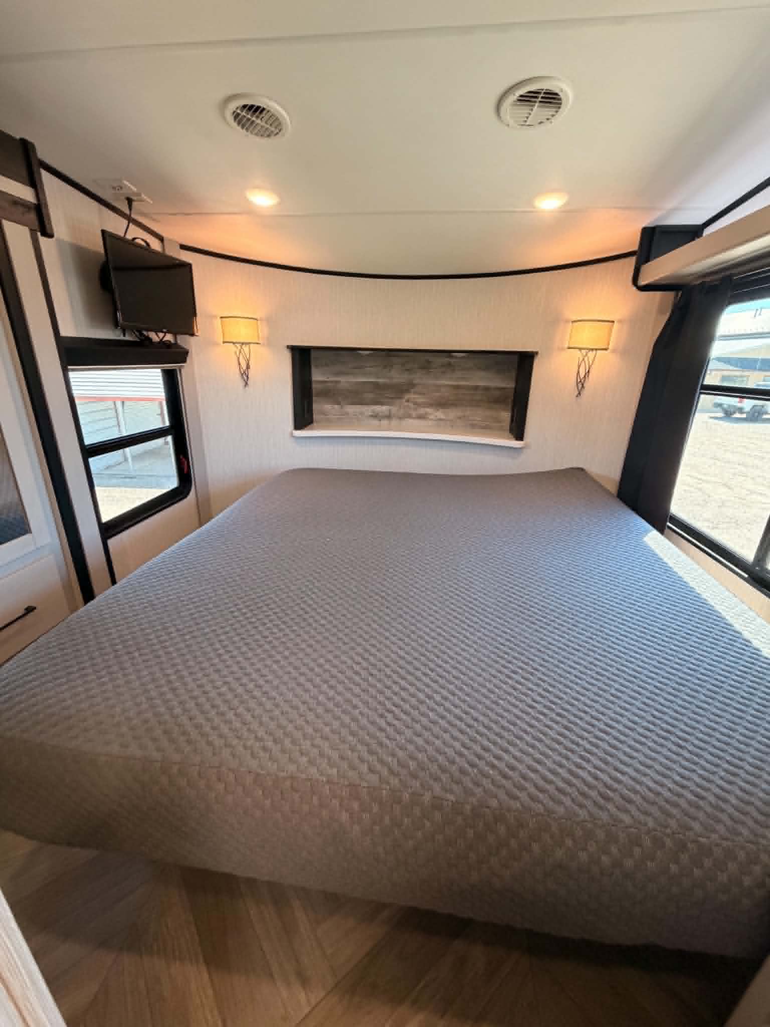 Comfortable RV rental bedroom featuring a large bed with grey quilt, wood floor, TV, windows, and wall sconces.. Heartland Cyclone 2022