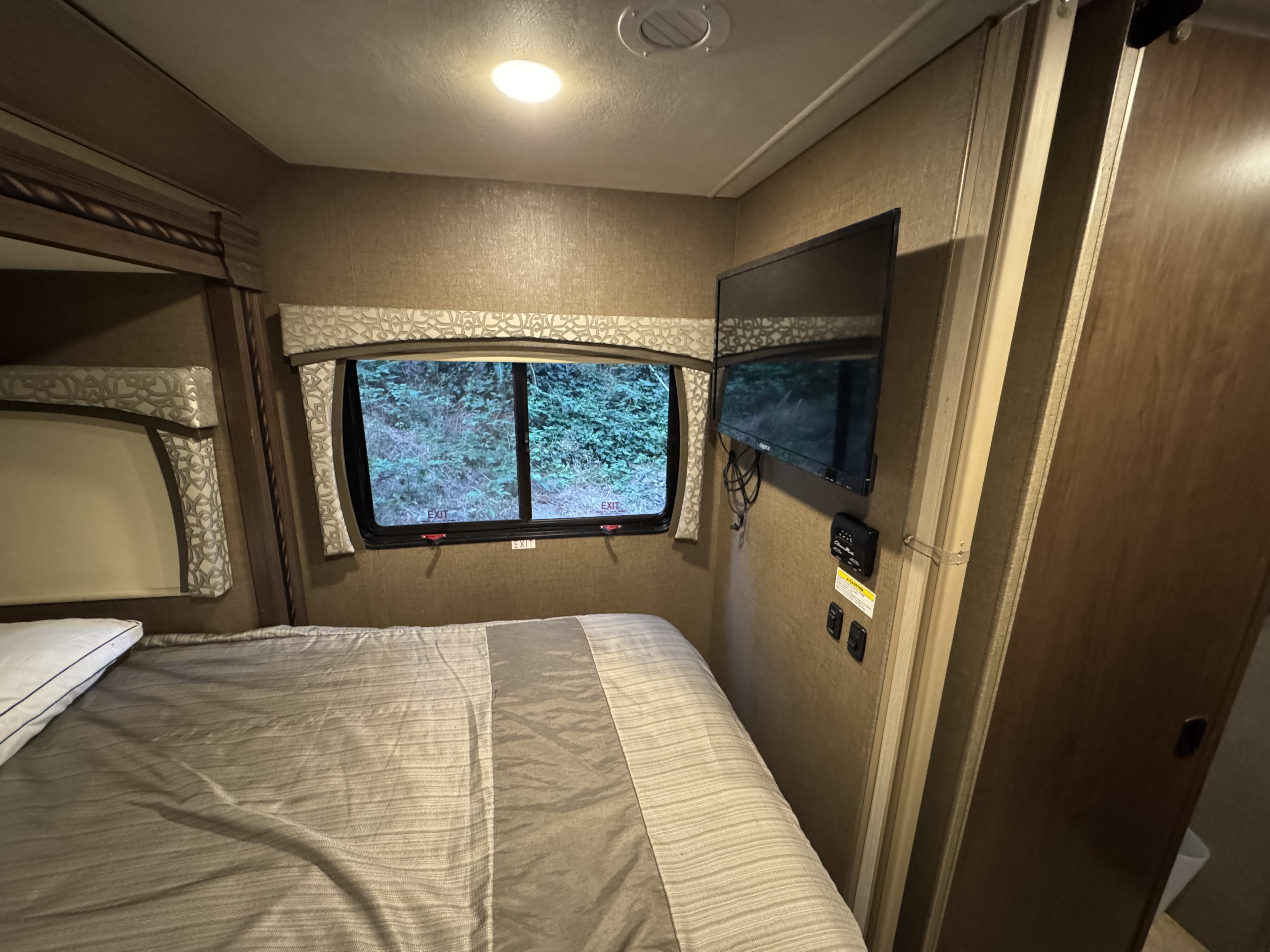 Cozy RV rental bedroom featuring a striped bed, window with nature views, mounted TV, and control panel. A compact, comfortable space.. Thor Chateau Sprinter 2018