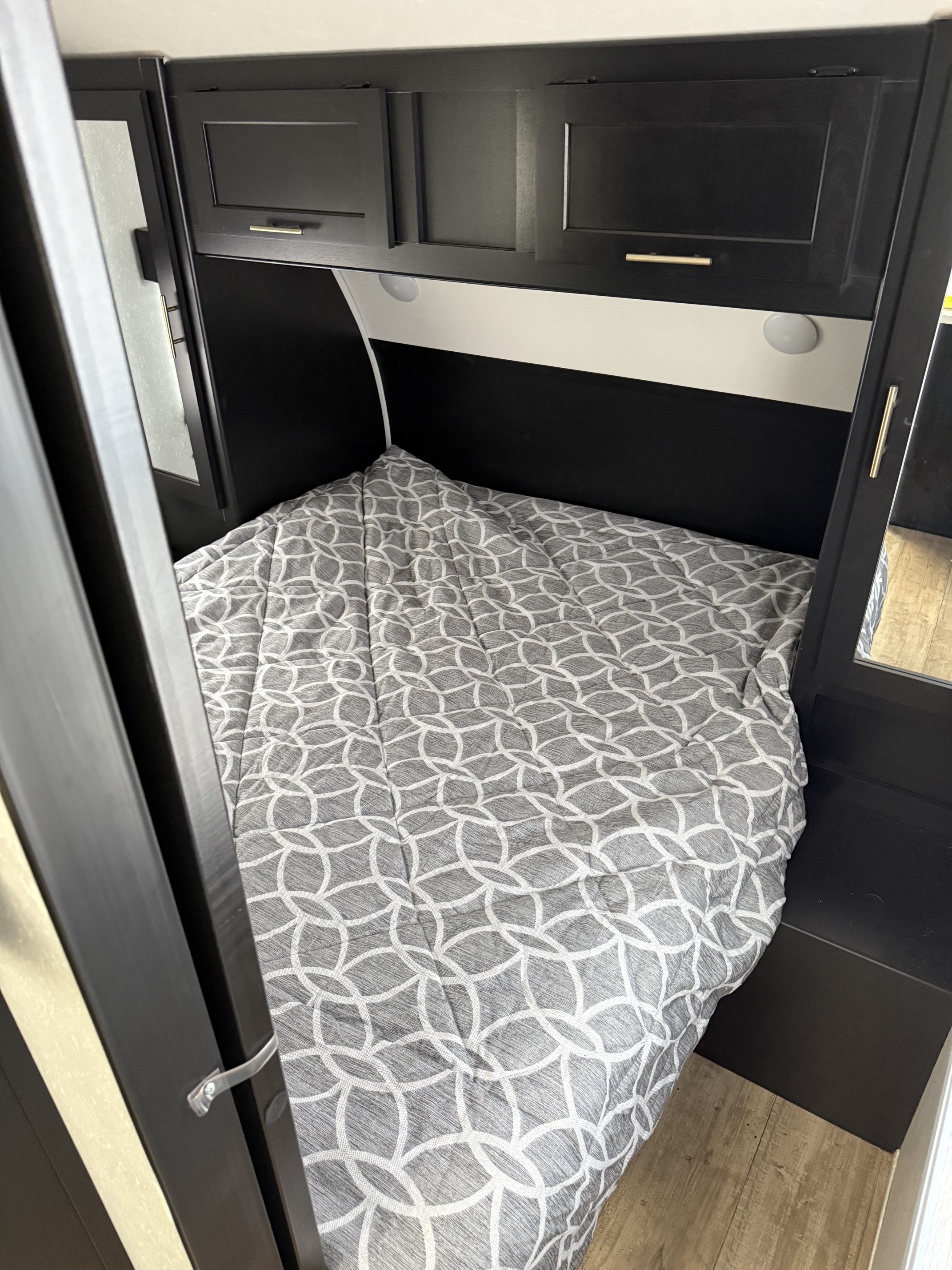 Cozy RV rental bedroom features a neatly made bed with a gray patterned comforter, overhead black cabinets, and reading lights.. Forest River Cherokee Grey Wolf 2022