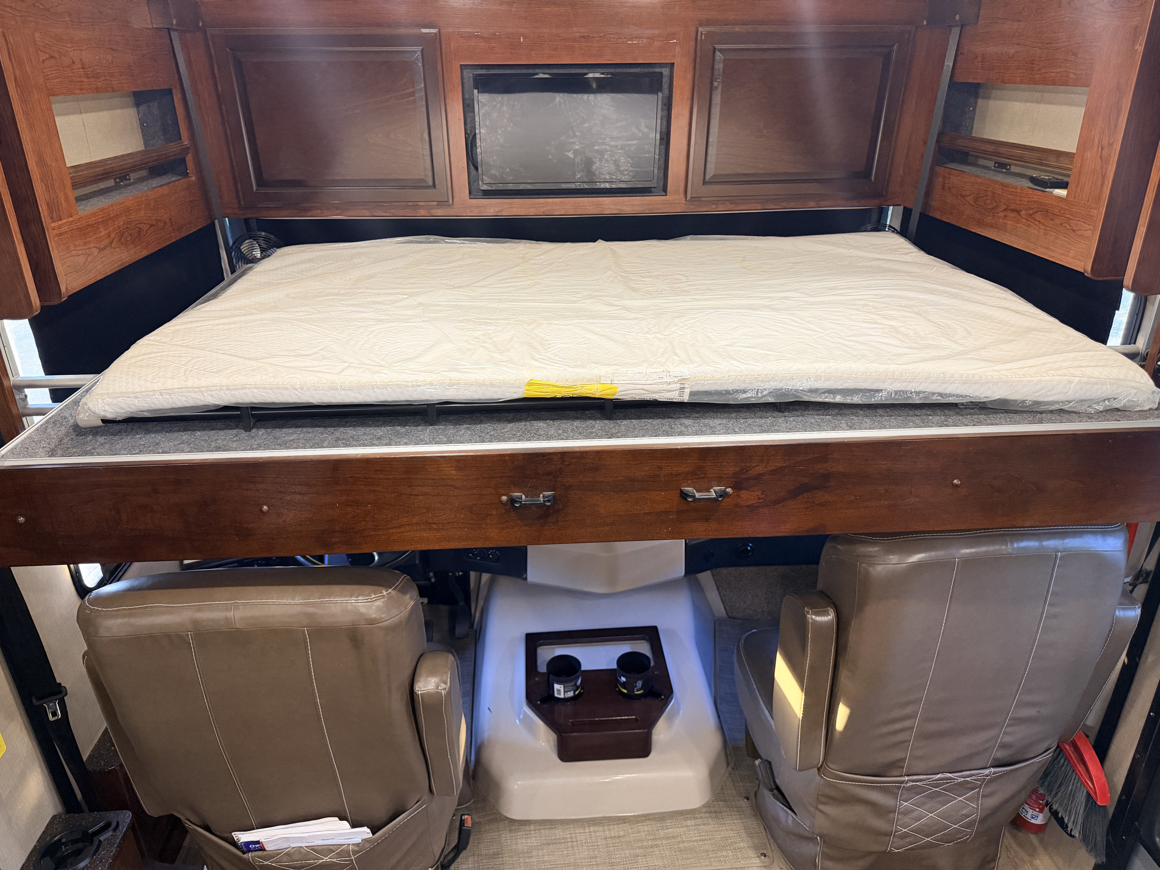 RV rental's cab-over bunk with new, plastic-wrapped mattress. Below are brown leather cockpit seats and overhead storage cabinets.. Fleetwood Bounder 2018