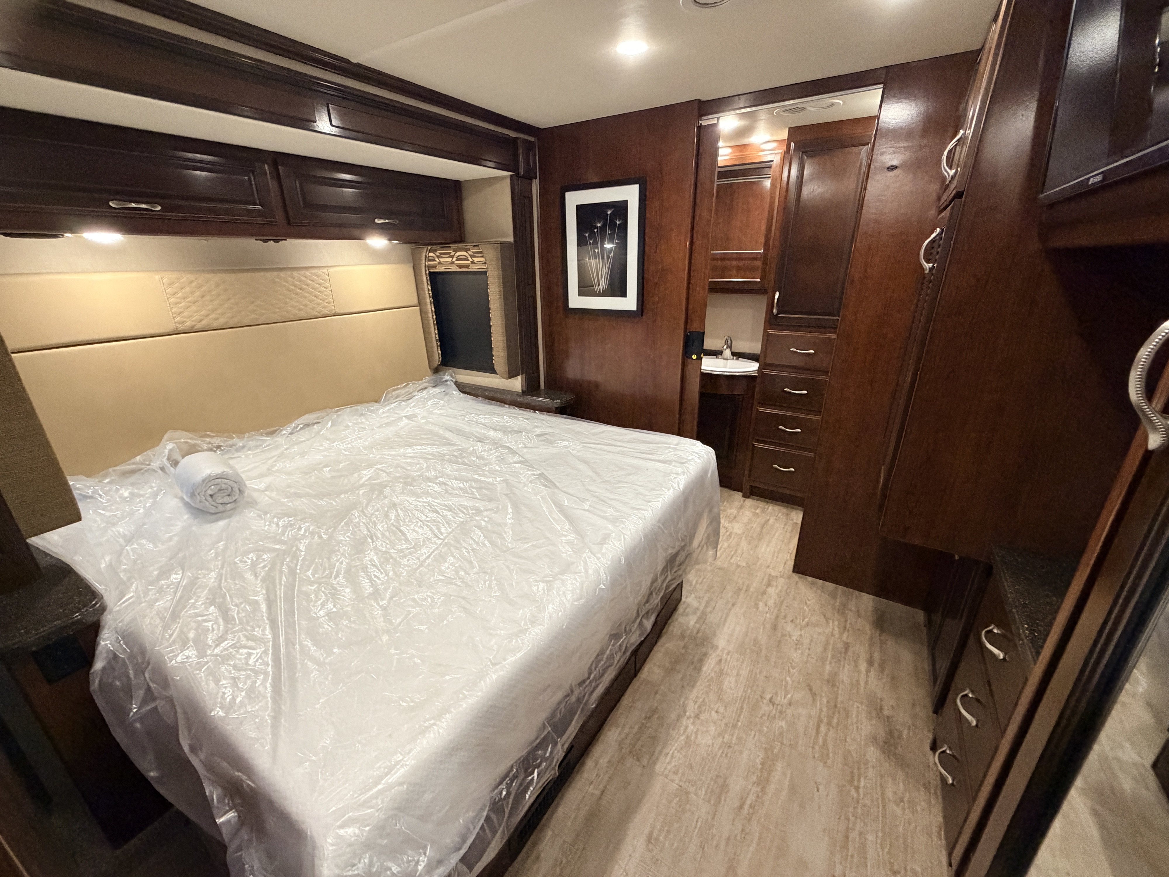 This RV rental bedroom features a plastic-covered bed, dark wood cabinets, and a compact vanity for a comfortable stay.. Fleetwood Bounder 2018