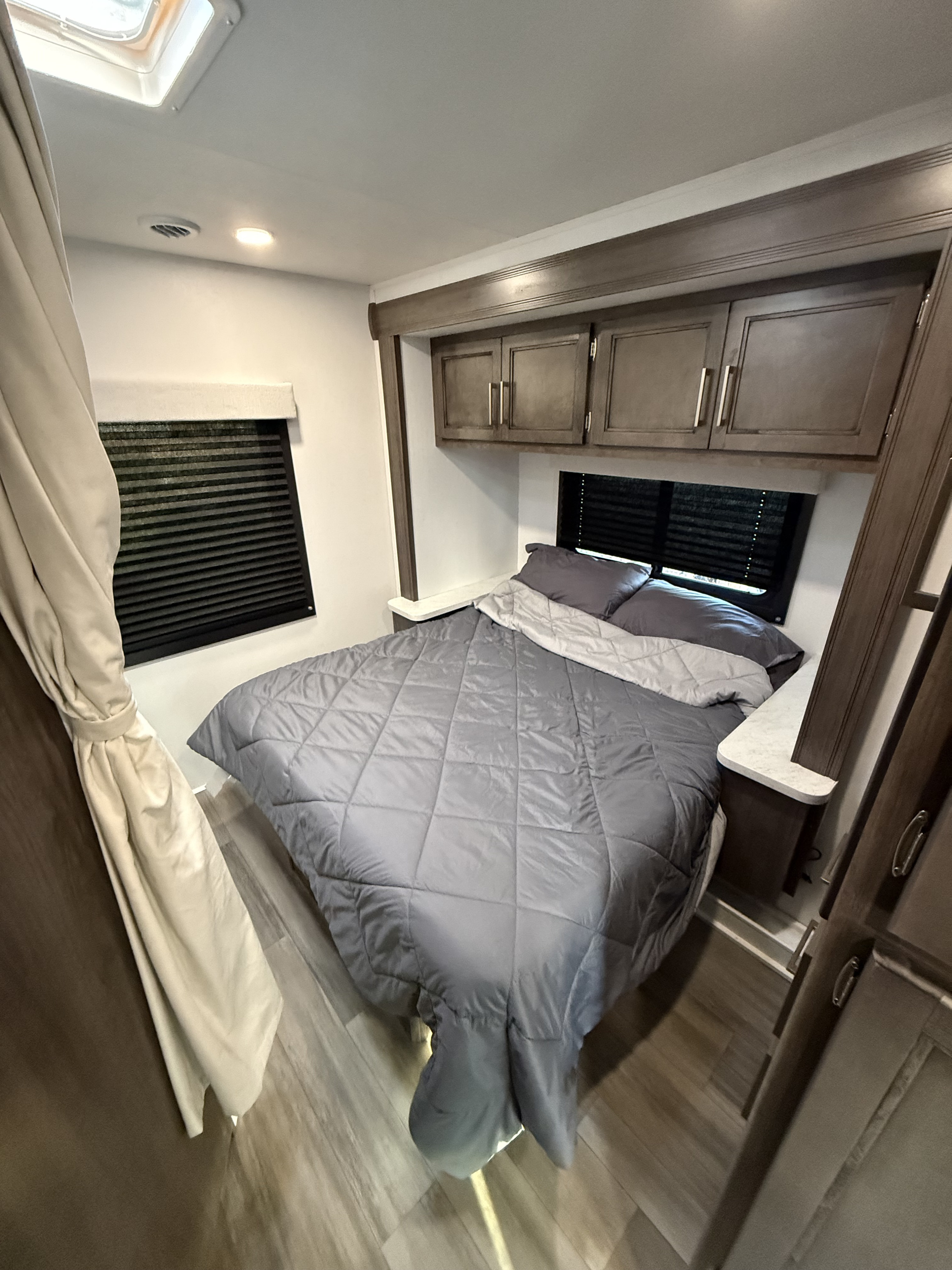 Cozy RV rental bedroom with a gray quilted bed, overhead storage cabinets, two windows with blinds, and a roof skylight.. Jayco Redhawk 22CF 2026
