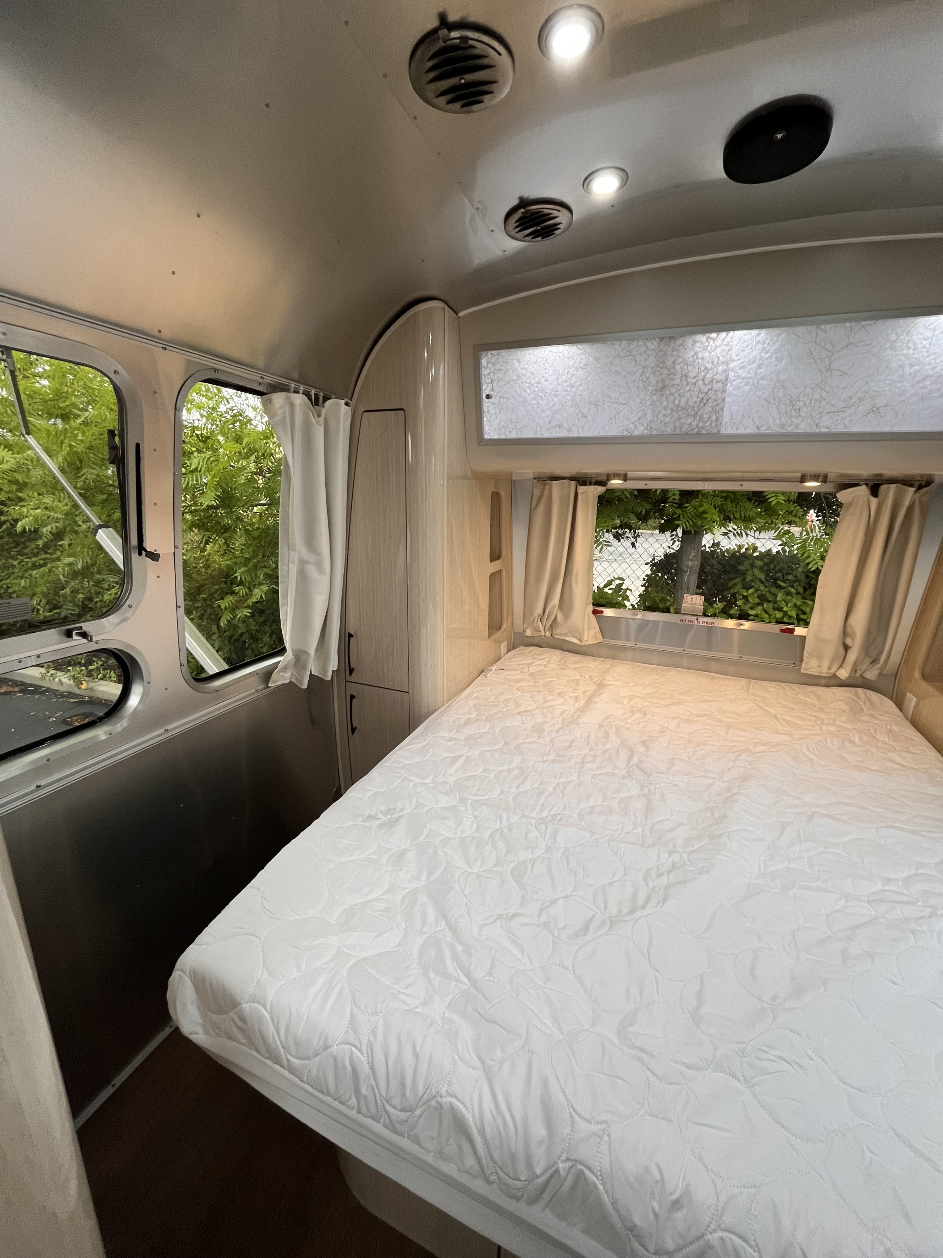 RV rental interior: A cozy bedroom with a white quilted bed, windows showing lush trees, and light wood storage.. Airstream International 2020