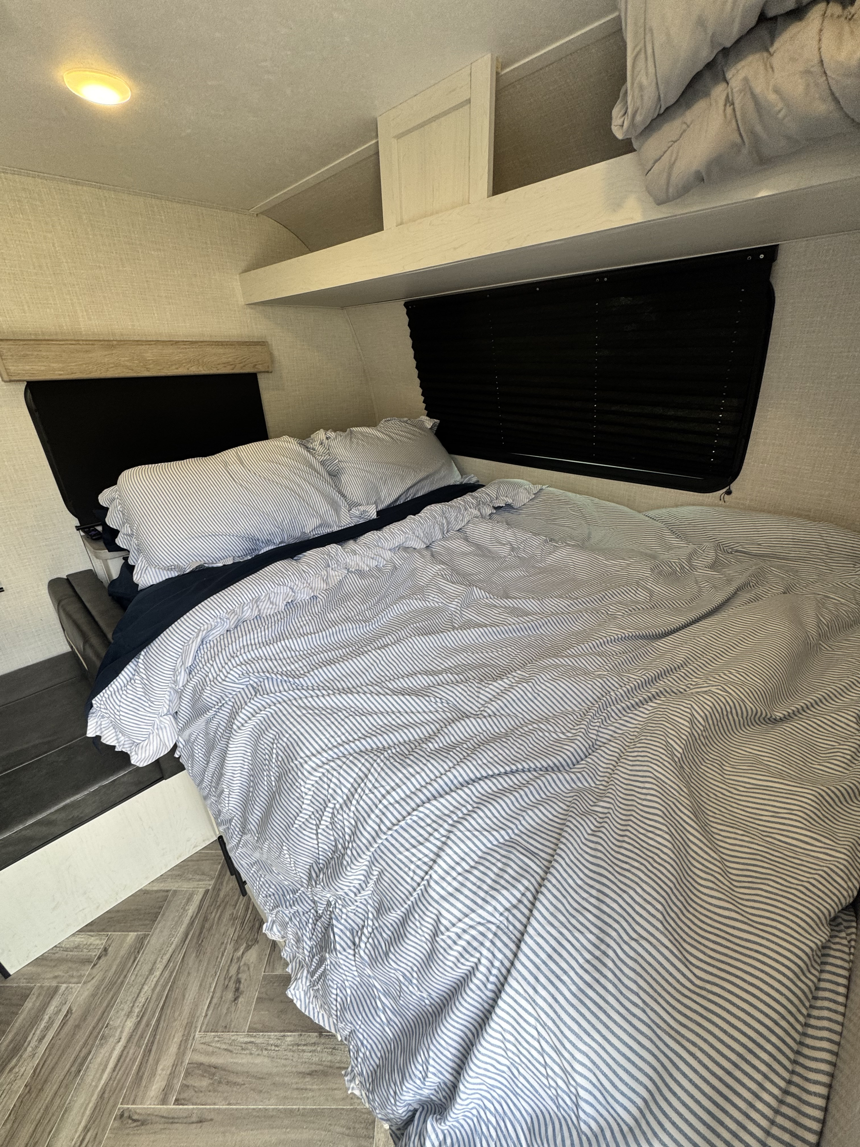 Cozy RV rental bedroom features a comfy bed with blue striped bedding, black headboard, overhead storage, and wood-look floor.. Dutchmen Coleman Rubicon 2023