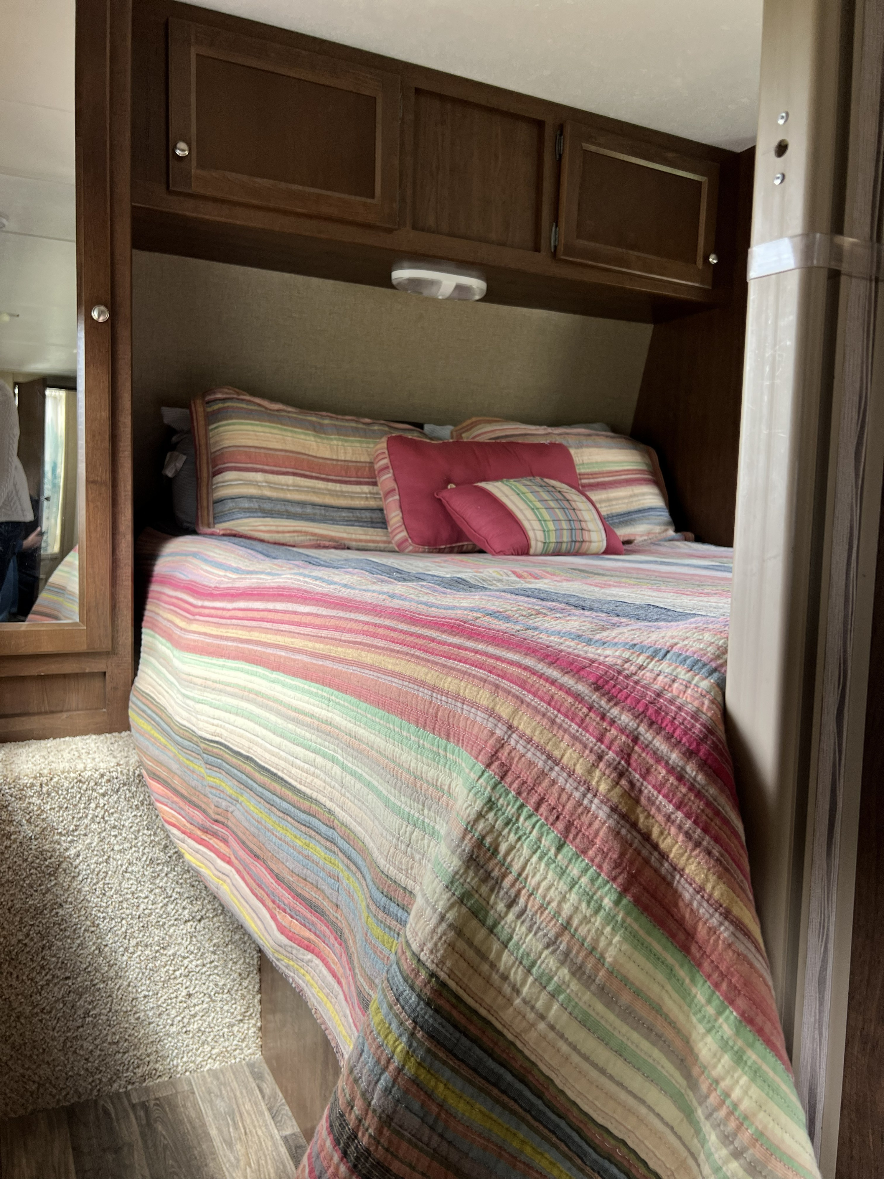 Cozy RV rental bedroom featuring a colorful striped quilt, pillows, overhead cabinets, and a mirror on the left.. Keystone Hideout 2015