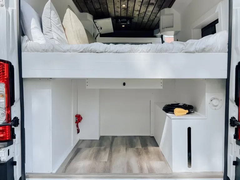 Inside an RV rental: White camper van interior with raised bed, lower cargo/storage area, and wooden ceiling.. Dodge B Van 2021