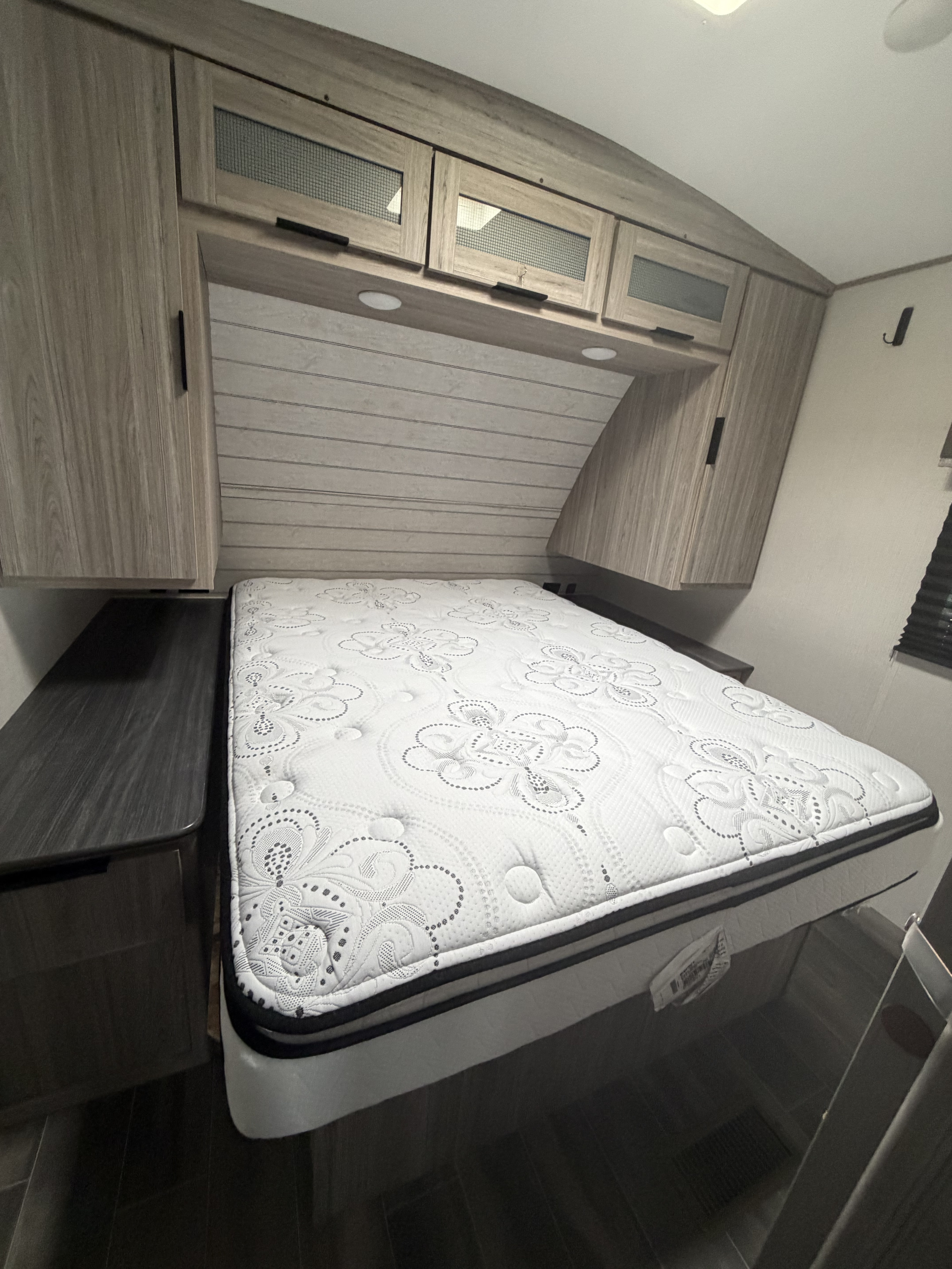 Inside an RV rental, a comfy bedroom features a patterned white mattress, light wood overhead cabinets, and dark bedside tables.. Keystone RV Passport GT 2021