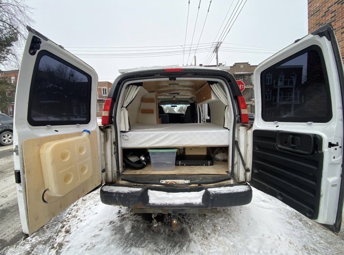 Open doors of an rv rental campervan, showing a bed, under-bed storage, and a water tank, ready for snowy vanlife trips.. Chevrolet Express 2500 2009