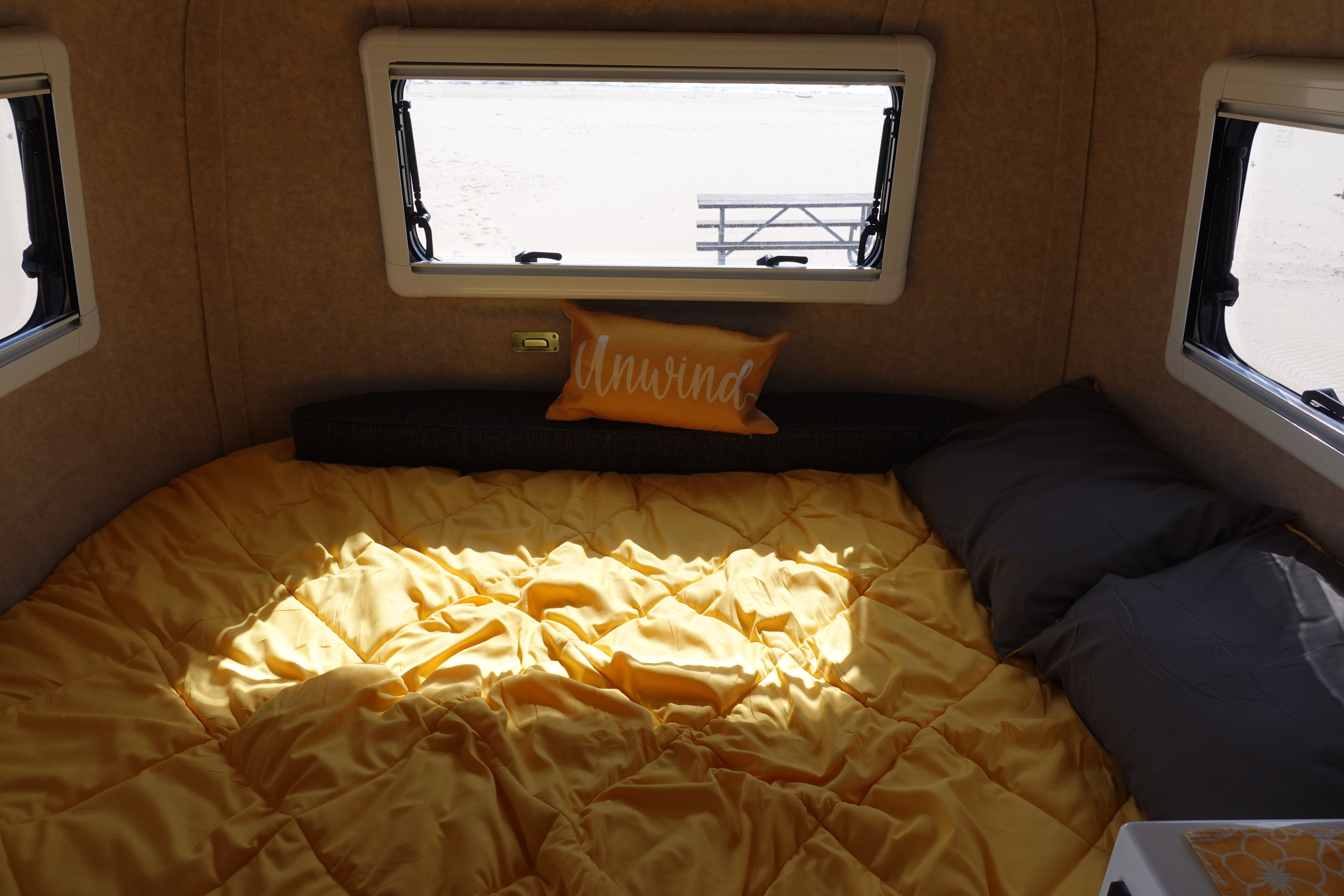 Cozy RV rental interior with a sunny bed, "Unwind" pillow, and a view of a beach and picnic table through the window.. Scamp Other 2026