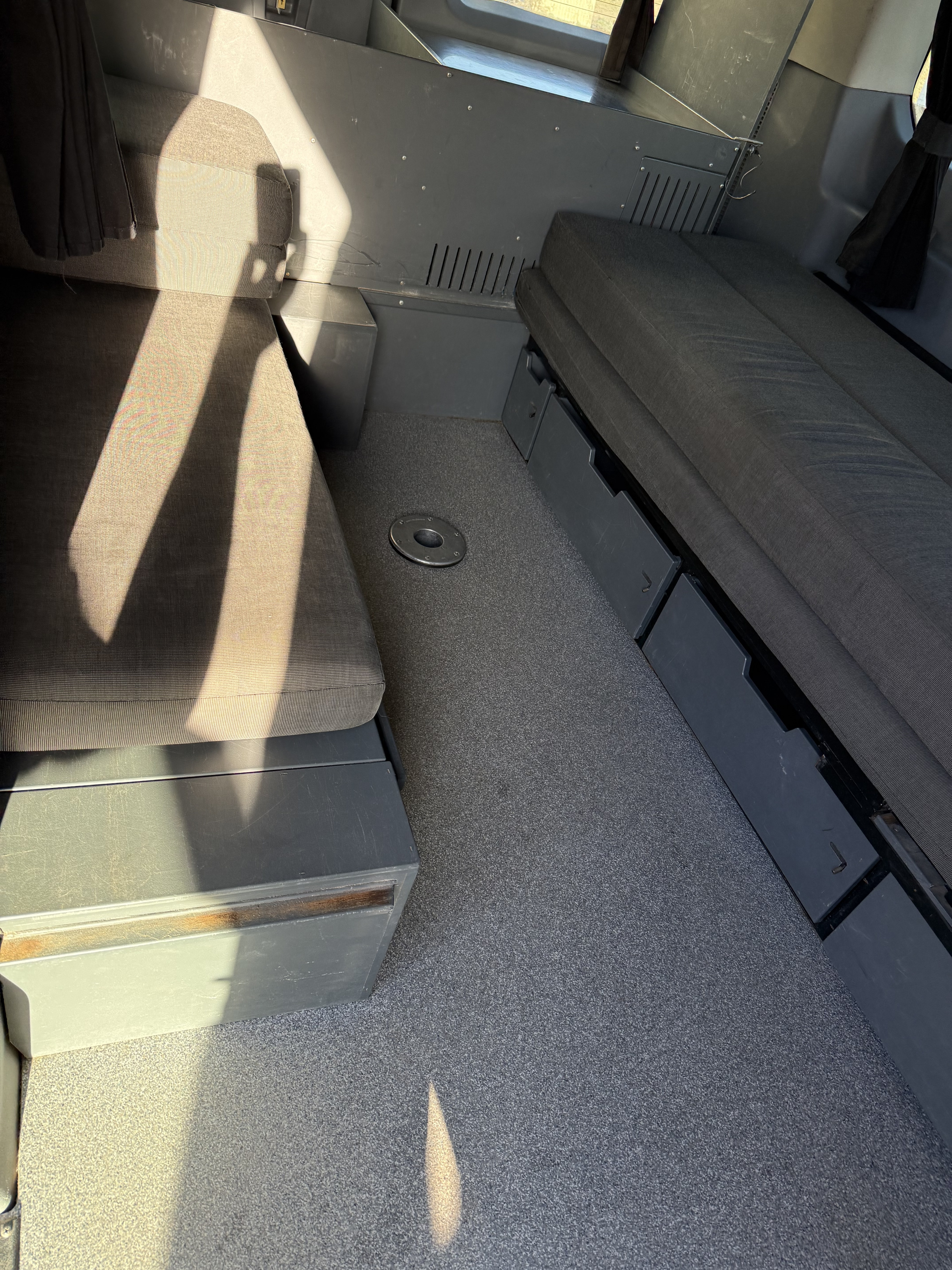 RV rental: Van interior showing cushioned benches, under-seat storage, table mount on grey carpet, sunlit.. Ford Transit 2017