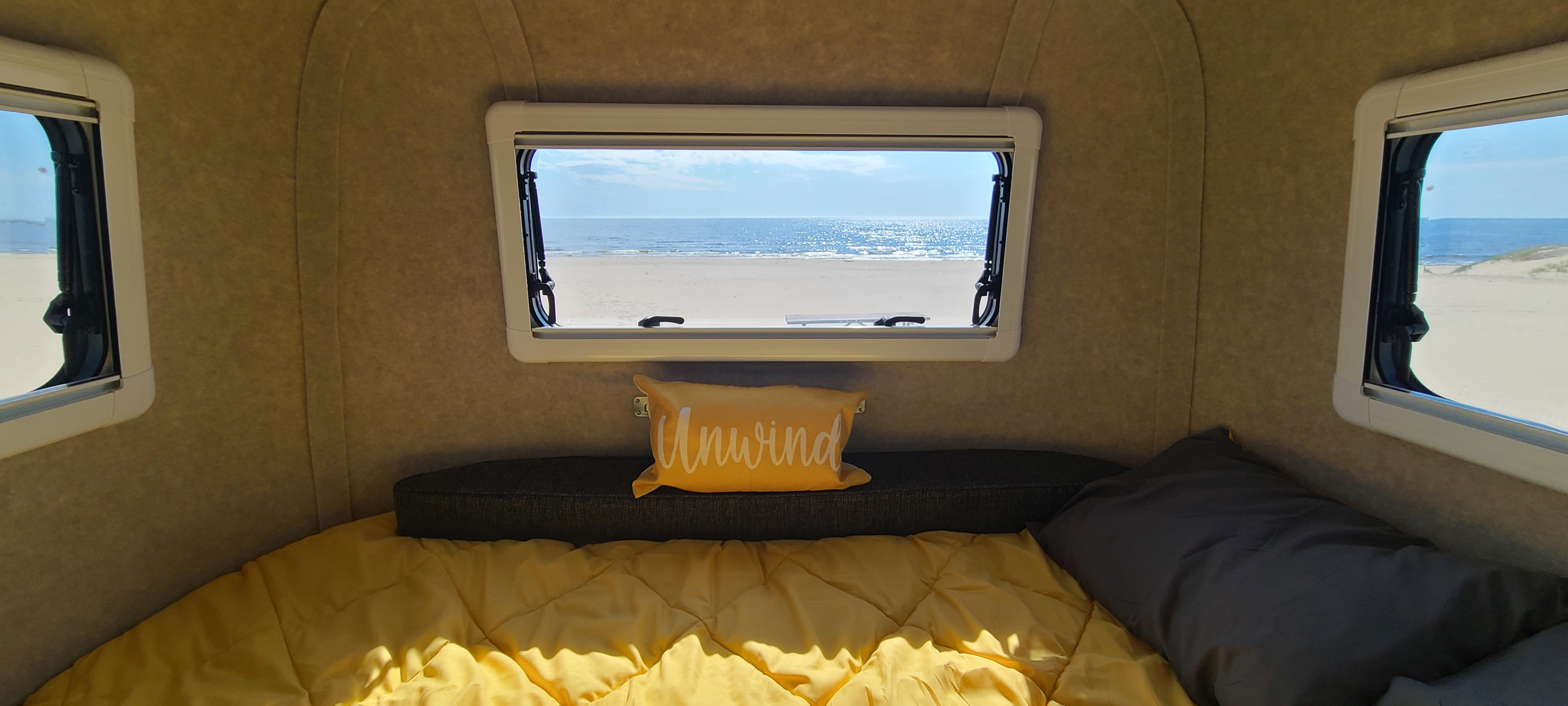 Cozy RV rental bedroom with a panoramic view of a sandy beach and sparkling ocean through the window. Yellow bedding invites relaxation.. Scamp Other 2026