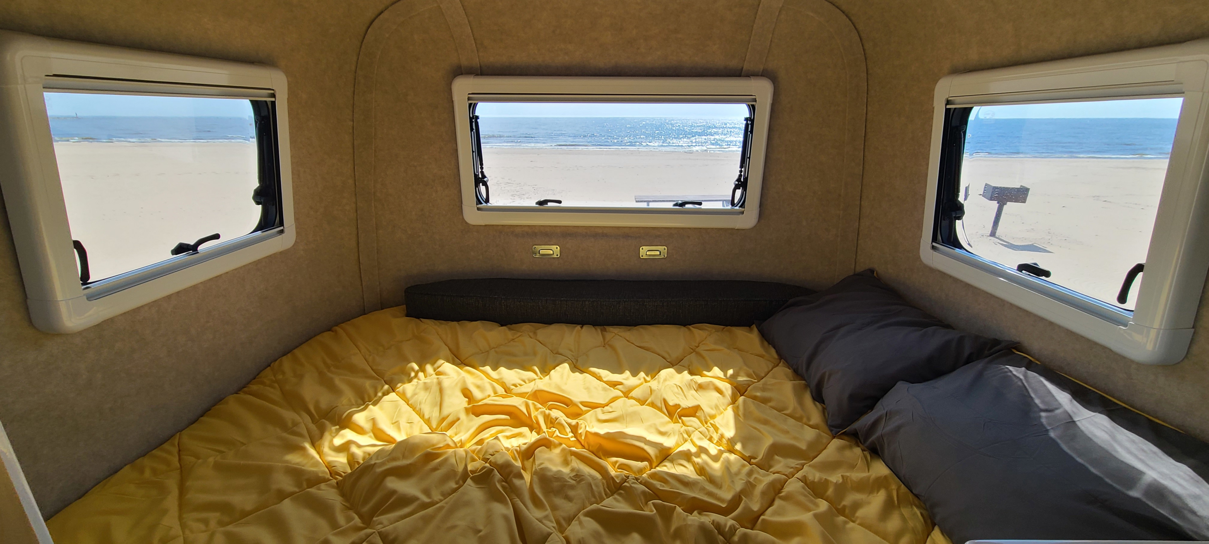 Inside a cozy RV rental: a bed with a yellow comforter and grey pillows. Three windows show a sunlit beach and ocean view.. Scamp Other 2026