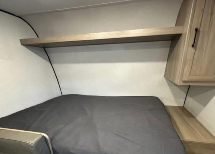 Cozy rv rental sleeping area featuring a bed, overhead shelf, and wooden storage cabinet for travel comfort.. Keystone RV Coleman 17B 2026