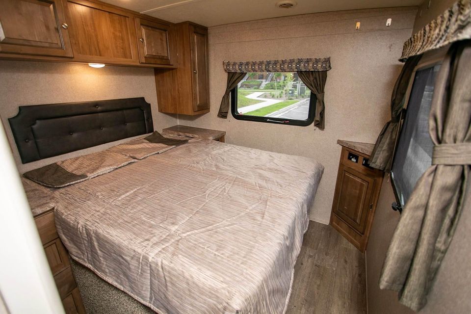 RV rental bedroom with a large bed, dark headboard, wooden overhead cabinets, and a window with patterned curtains.. Forest River Rockwood Mini Lite 2018