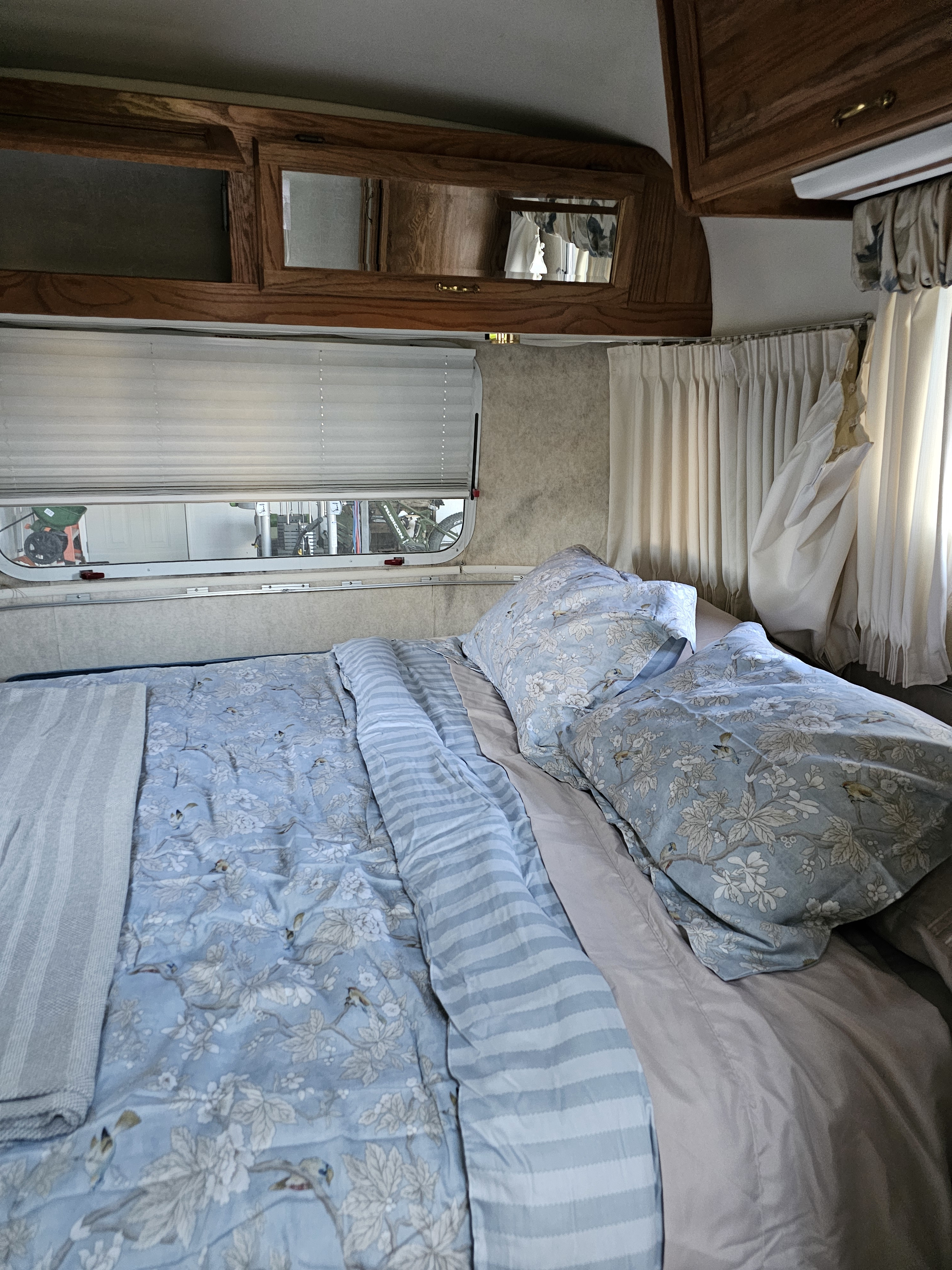 Cozy RV rental bedroom featuring a bed with blue floral bedding and striped comforter, window blinds, and wooden overhead cabinets.. Airstream Excella 2001