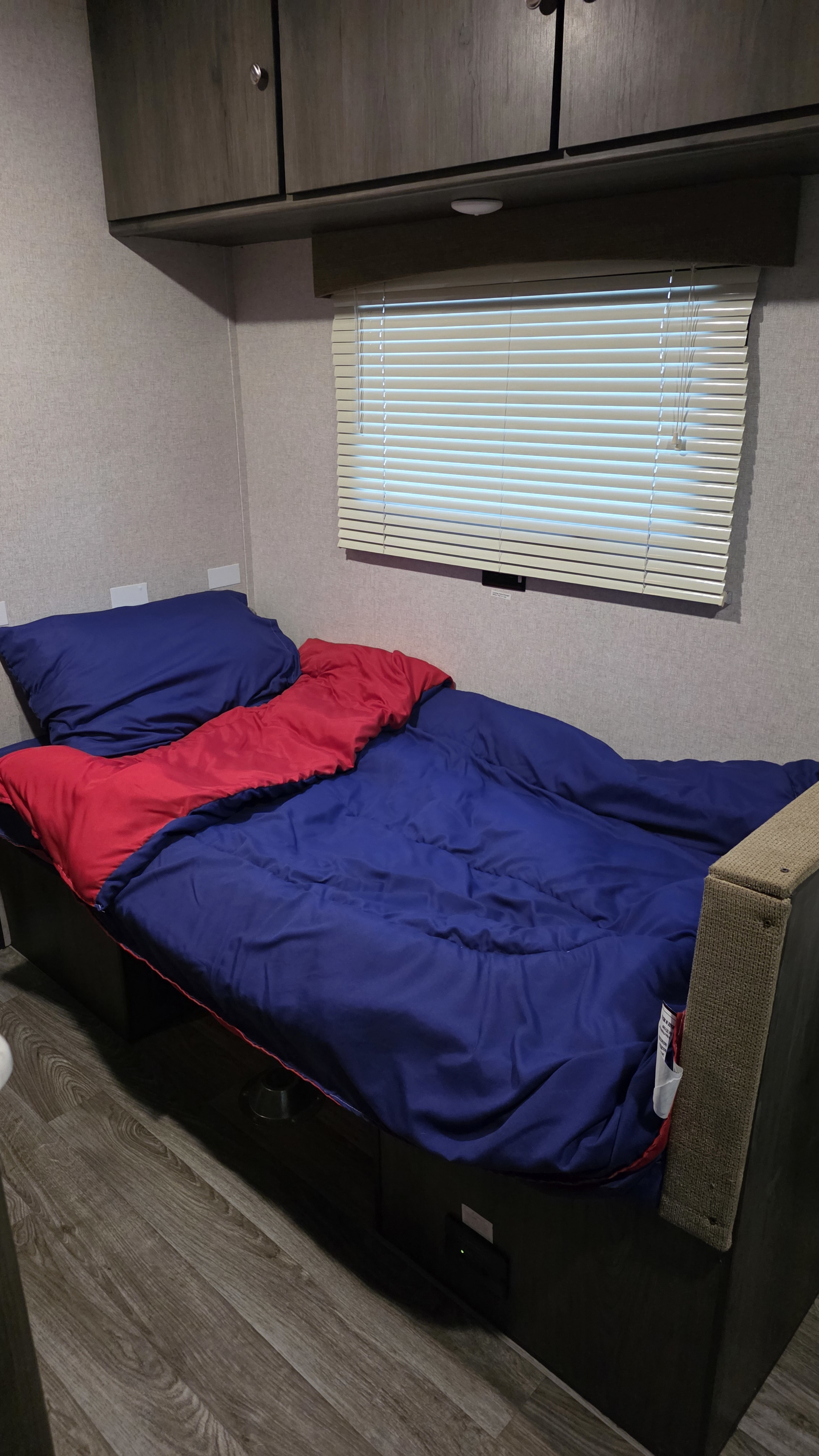 Compact RV rental bedroom featuring a blue and red bed, overhead storage, and a window with white blinds.. Clipper Cadet 2020