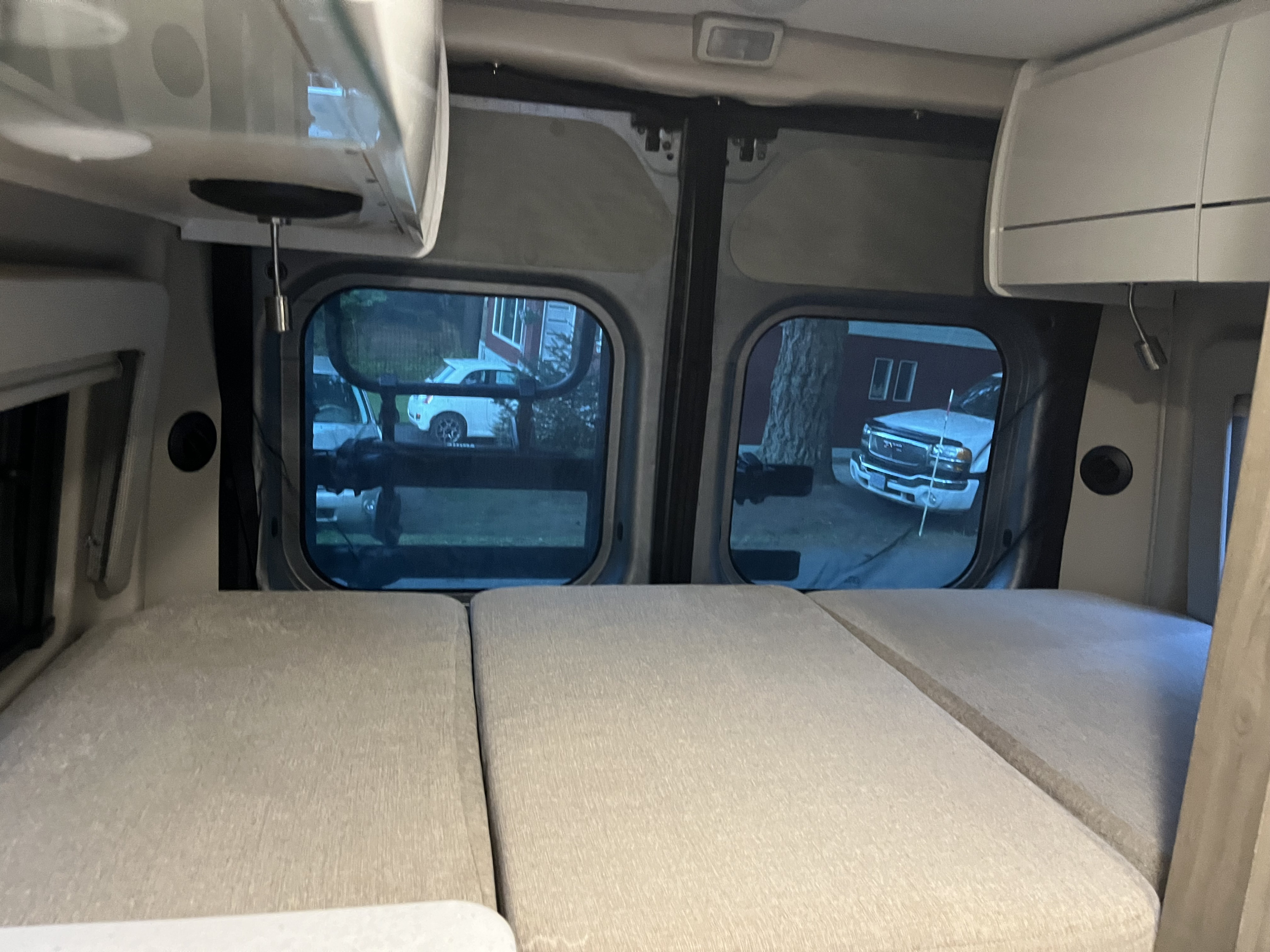 RV rental camper van interior: a bed, rear windows viewing a car and truck outside, and white overhead cabinets.. Thor Rize 2023