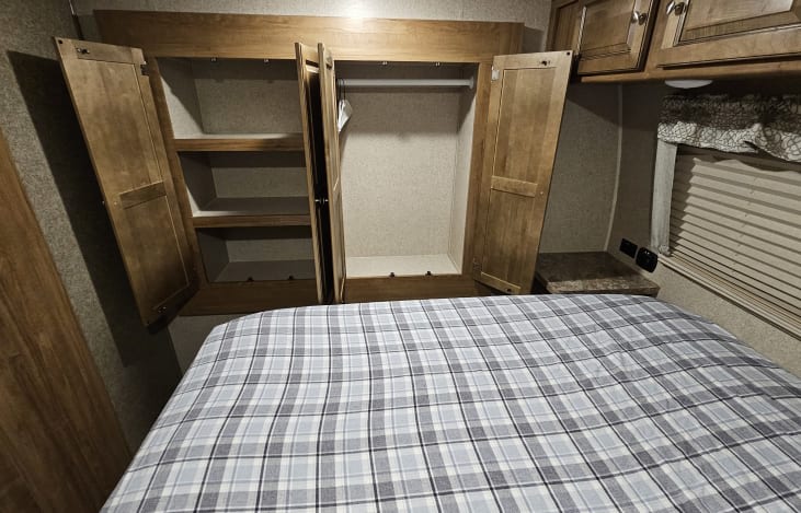 RV rental bedroom with plaid bed, open wooden cabinets, and wardrobe storage. Perfect for your travels.. Forest River Rockwood Ultra Lite 2019