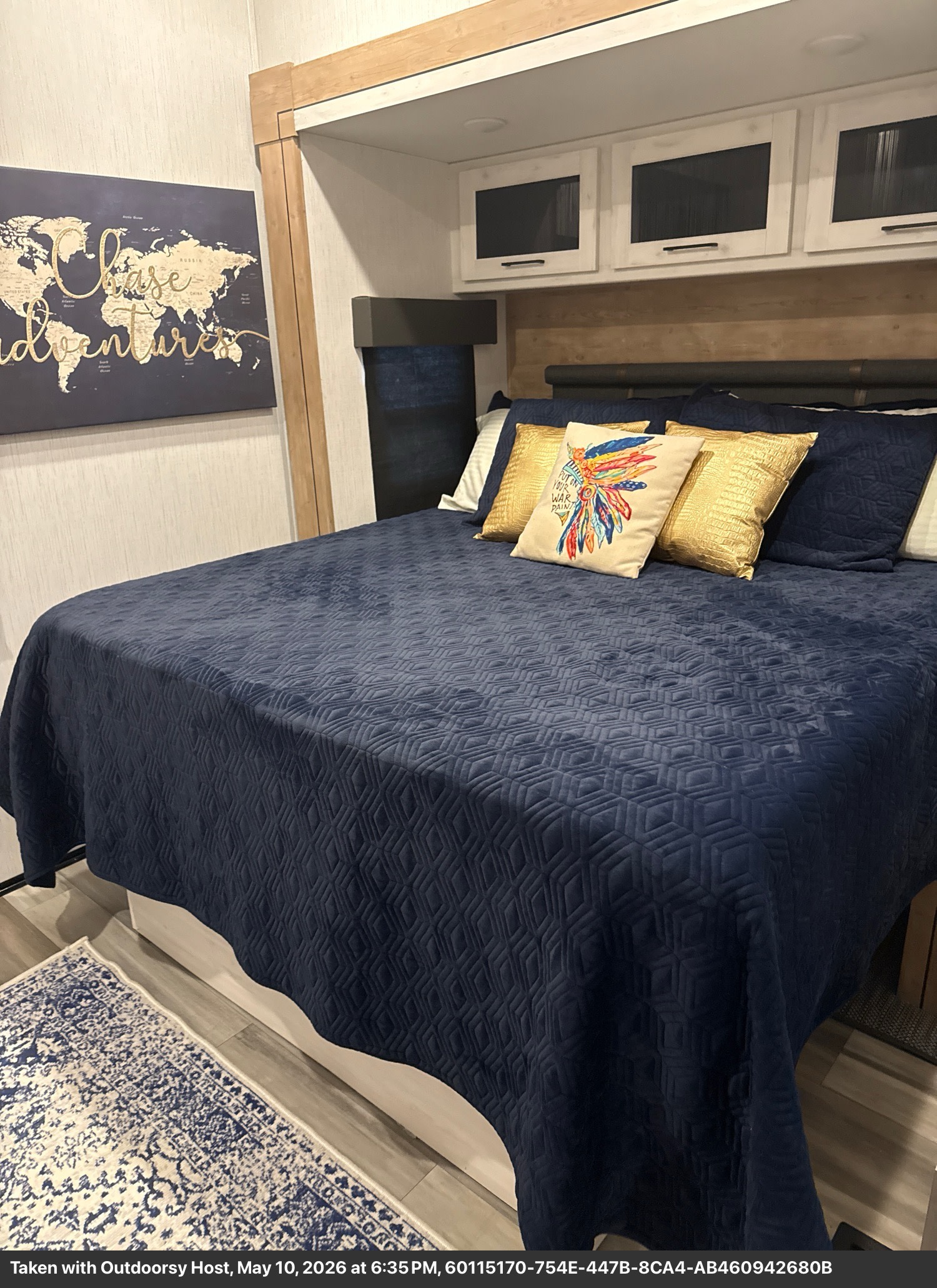 Cozy RV rental bedroom featuring a dark blue quilted bedspread, decorative pillows, map art, and convenient overhead storage.. Heartland RVs Milestone 2023