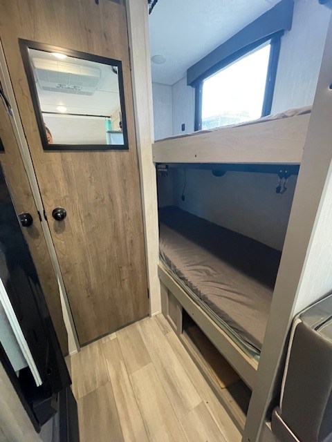 Inside an RV rental: a wooden door with a mirror on the left, bunk beds with a window above on the right. Cozy and practical.. Coachmen Clipper 2025