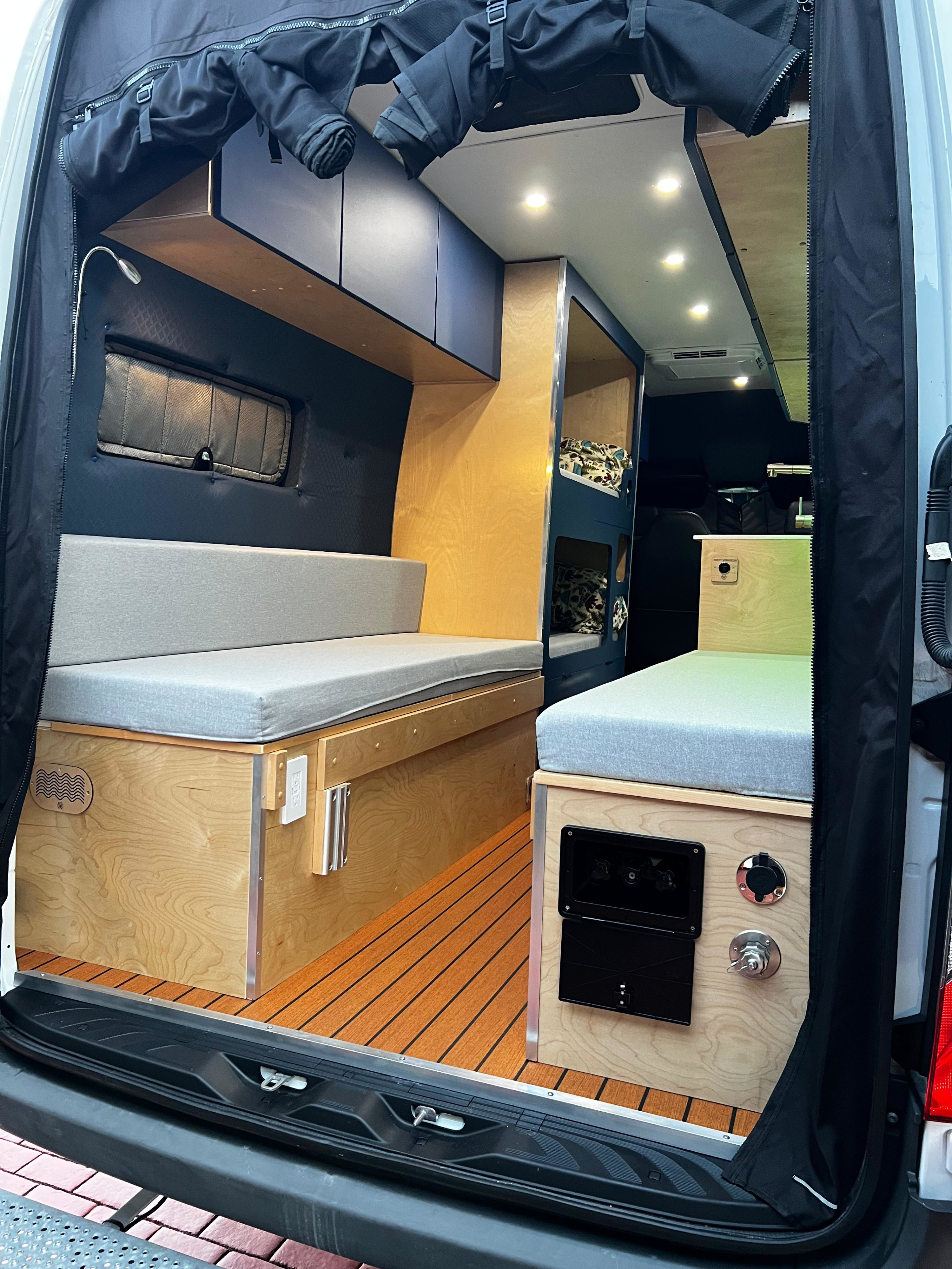 This inviting RV rental's camper van interior features light wood, grey cushions, bunk beds, and a slatted floor, seen from the rear.. Mercedes-Benz Sprinter 2023