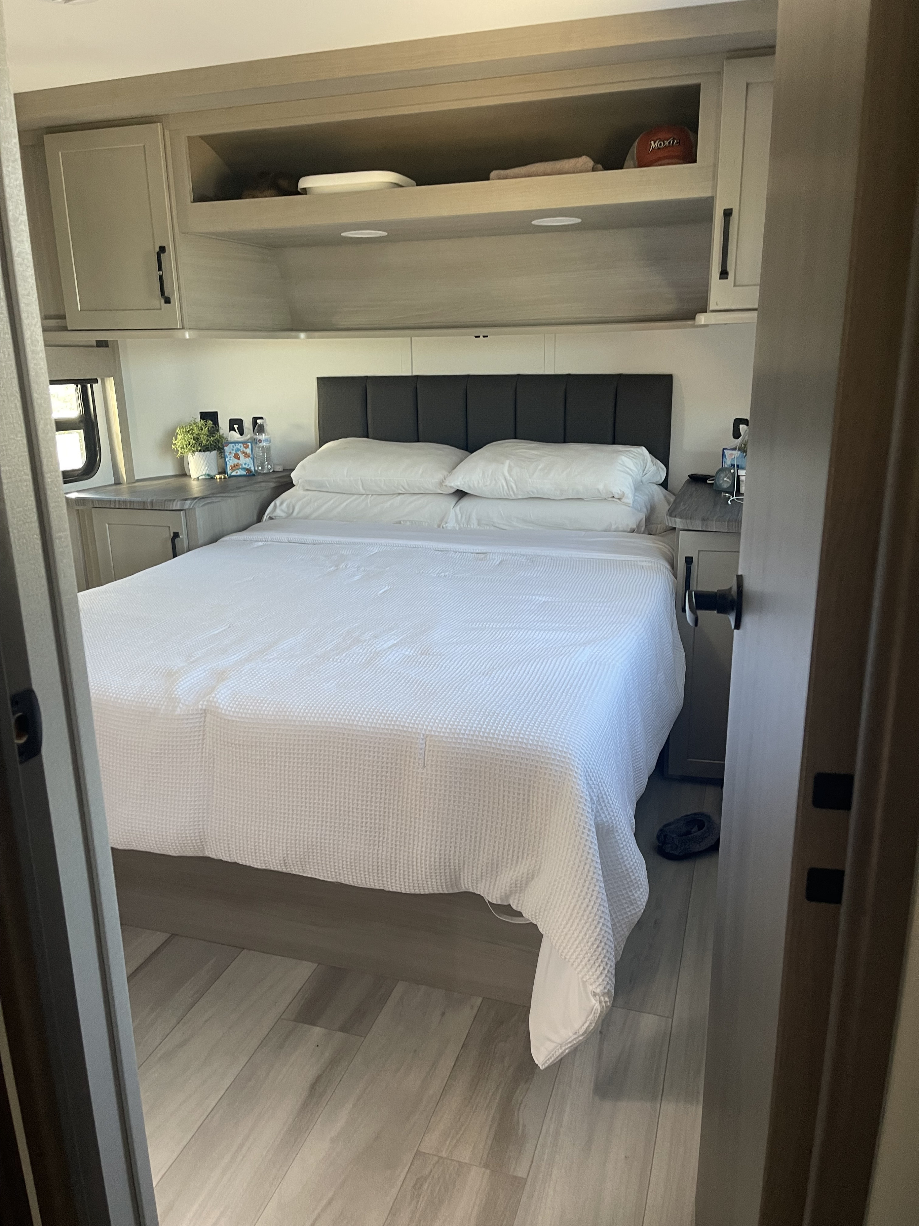 Inside an rv rental: a cozy bedroom with a white bed, dark headboard, light wood cabinets, and shelving.. Grand Design Momentum M-Class Toy Hauler 2025