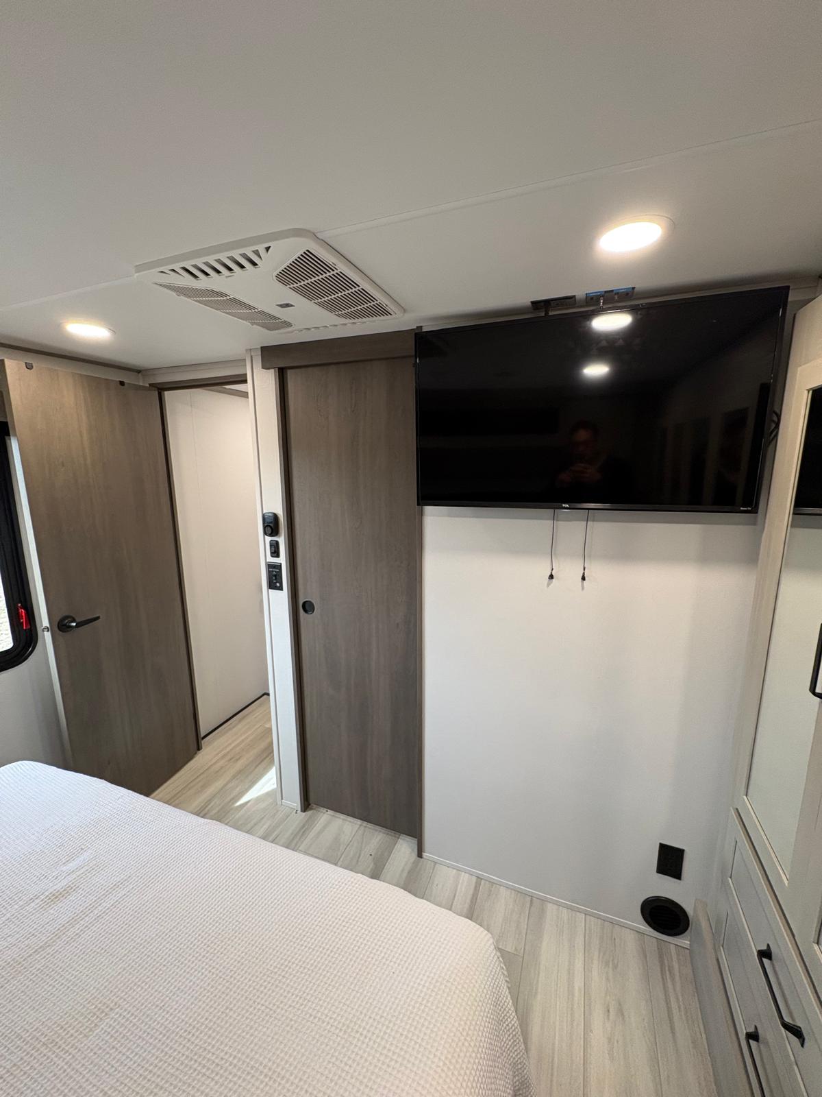 Inside a modern RV rental bedroom featuring a flat-screen TV, ceiling AC, light flooring, and comfortable bed.. Grand Design Momentum M-Class Toy Hauler 2025