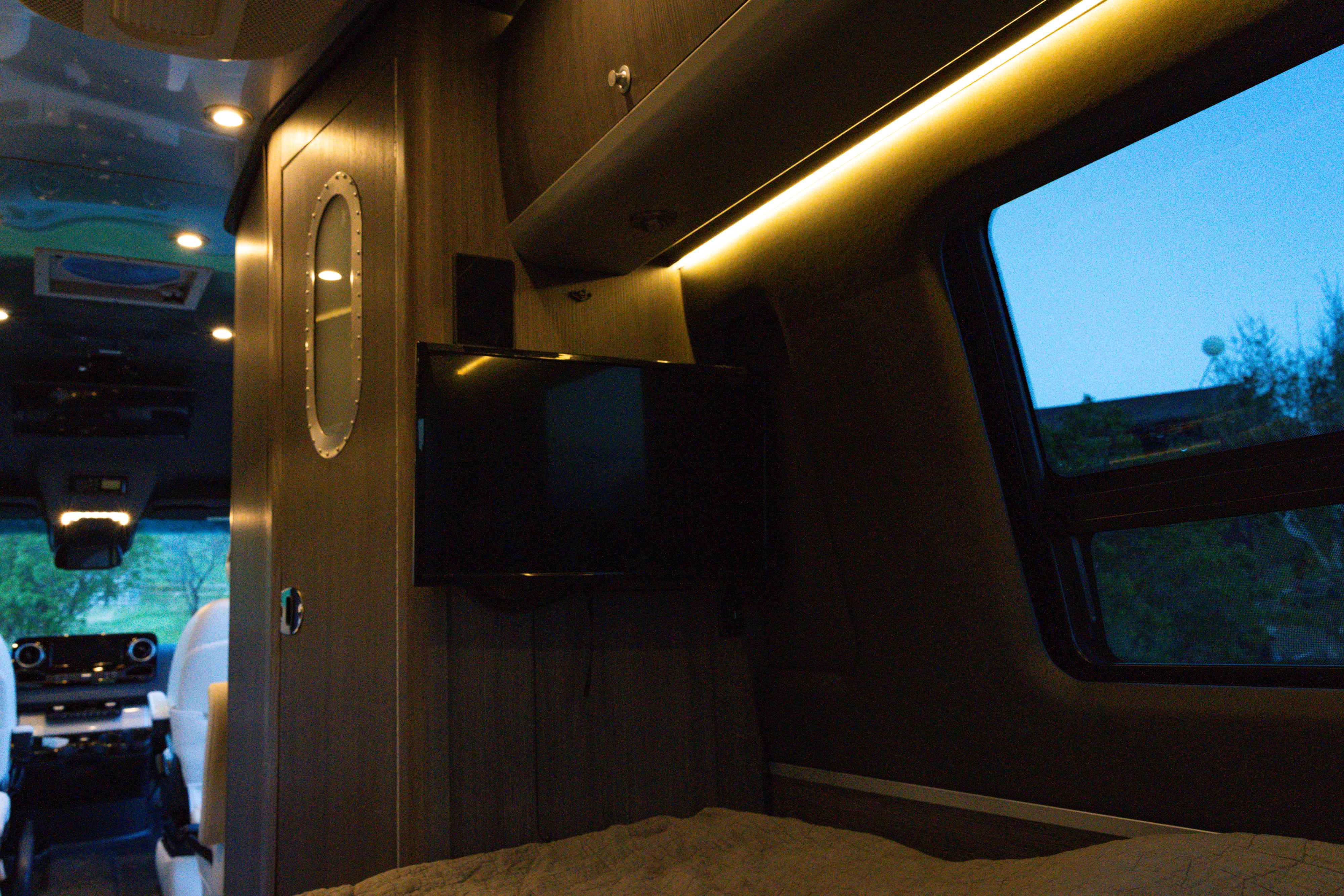 Interior of an **rv rental**. From the bed, a TV, overhead lighting, and a window viewing a twilight landscape are visible.. Airstream Interstate 2022