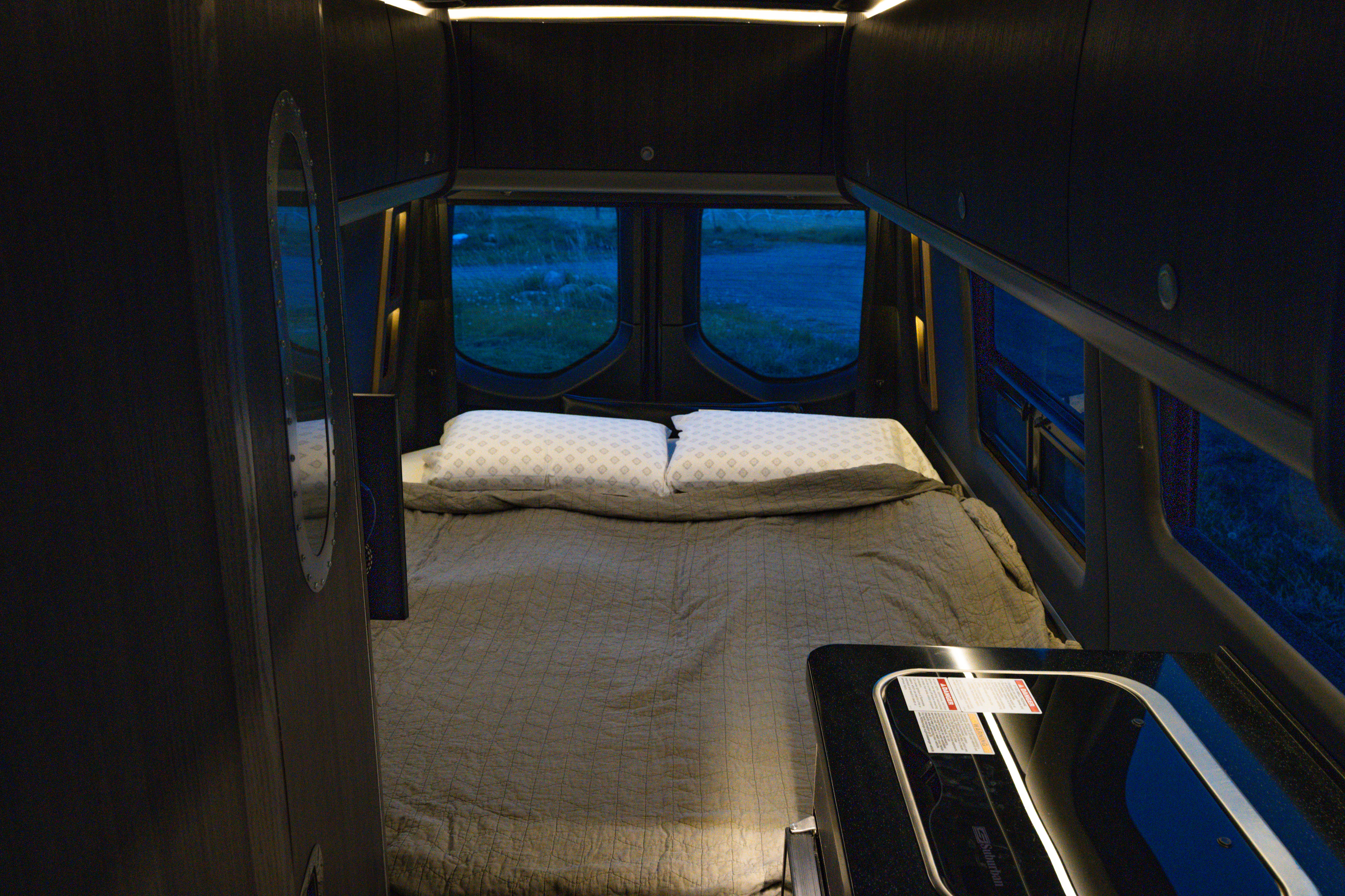 Cozy RV rental interior at night: bed, mirror, and kitchen counter visible. Windows show a dark outdoor landscape.. Airstream Interstate 2022