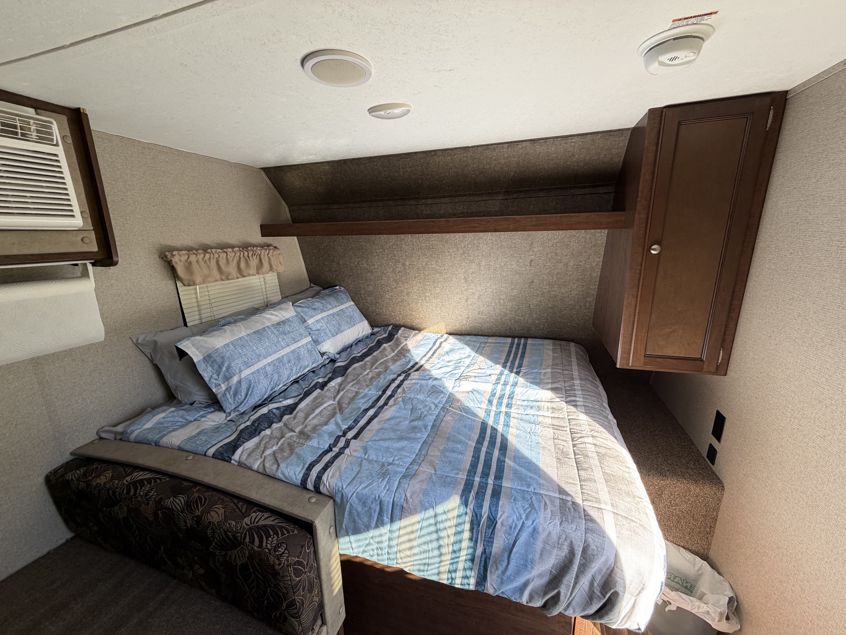 Inside an rv rental, a bedroom with striped bedding, AC, overhead storage, and a cabinet. Sunlight streams across the bed.. Keystone RV Hideout LHS 2018